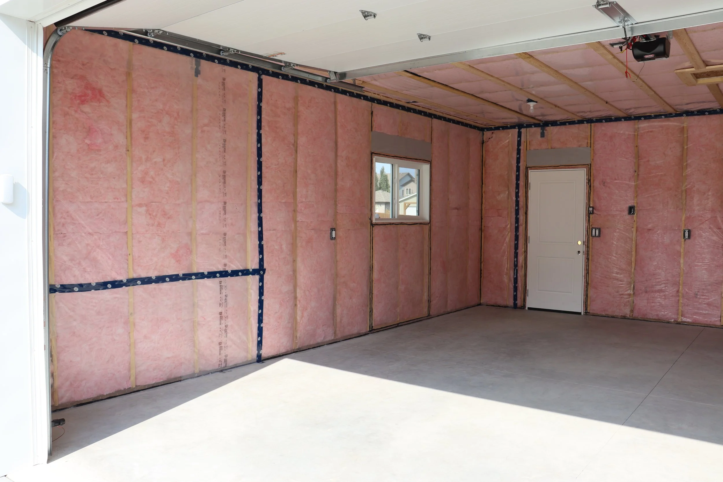An unfinished garage with pink insulation on the walls, a small window, a white door, and an open garage door with sunlight streaming in.