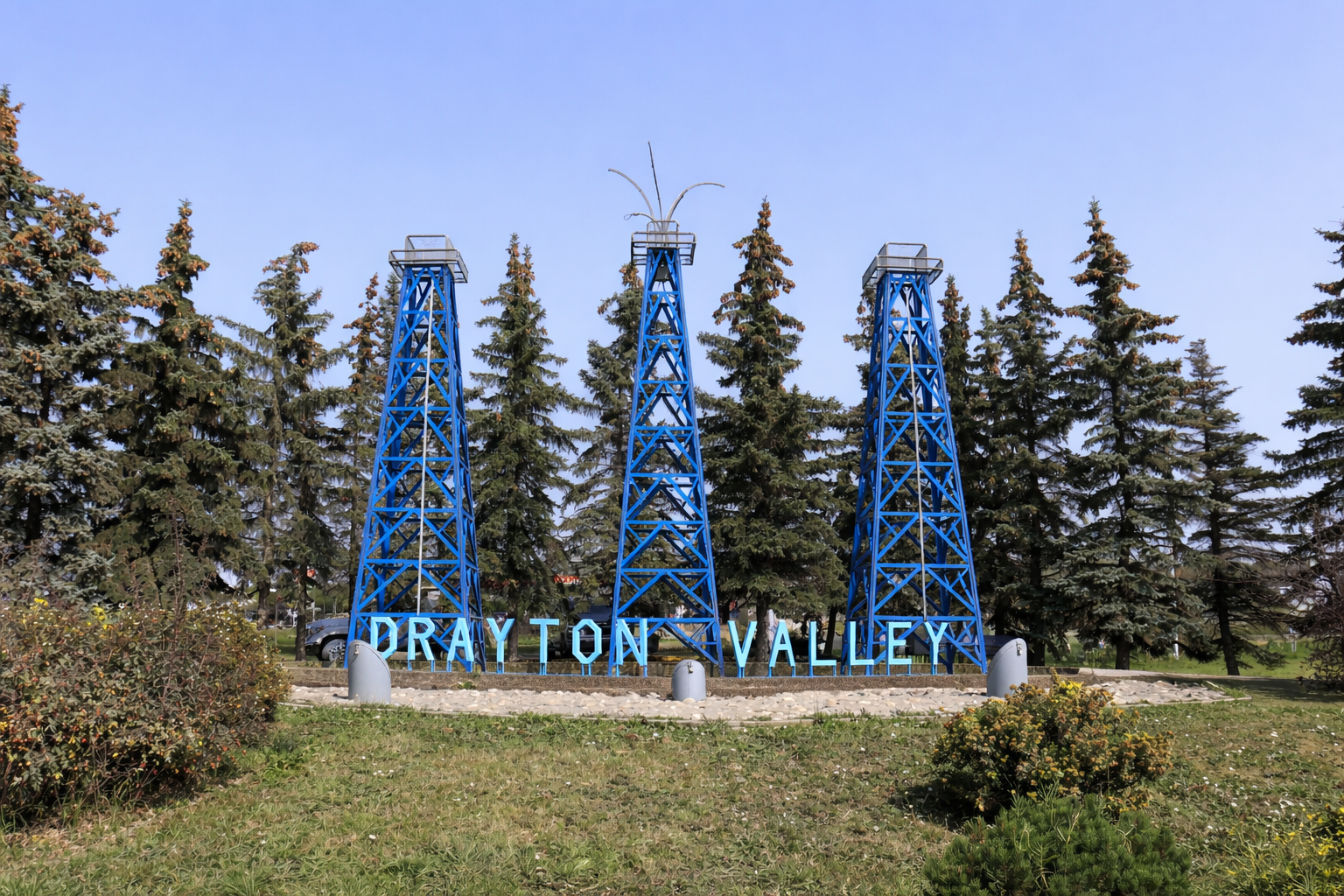 Sign for Drayton Valley featuring three blue oil derricks and surrounded by trees and grass.