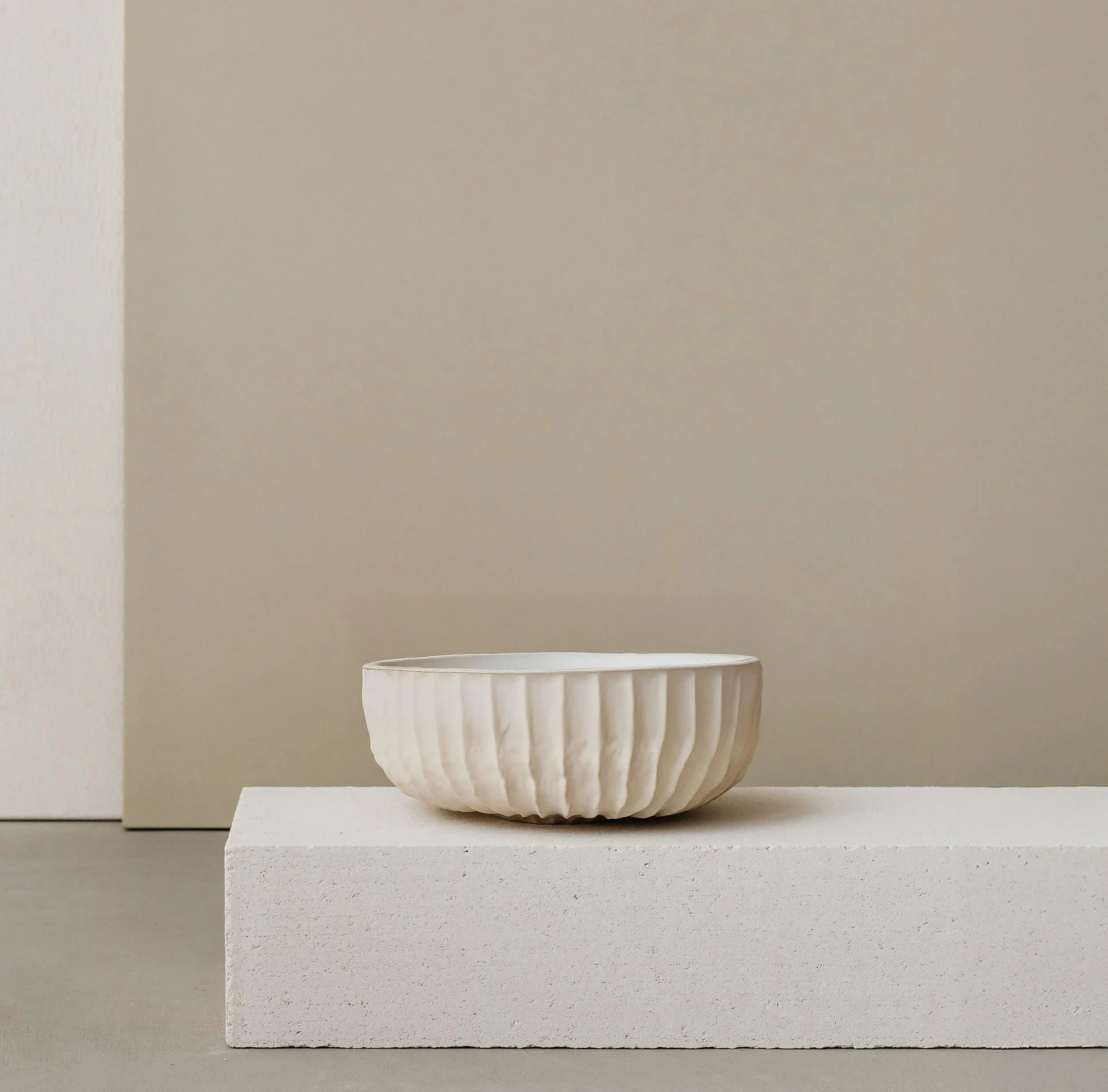 Handmade ceramic bowl with a ribbed texture, displayed on a minimalist off-white plinth against a soft beige background.