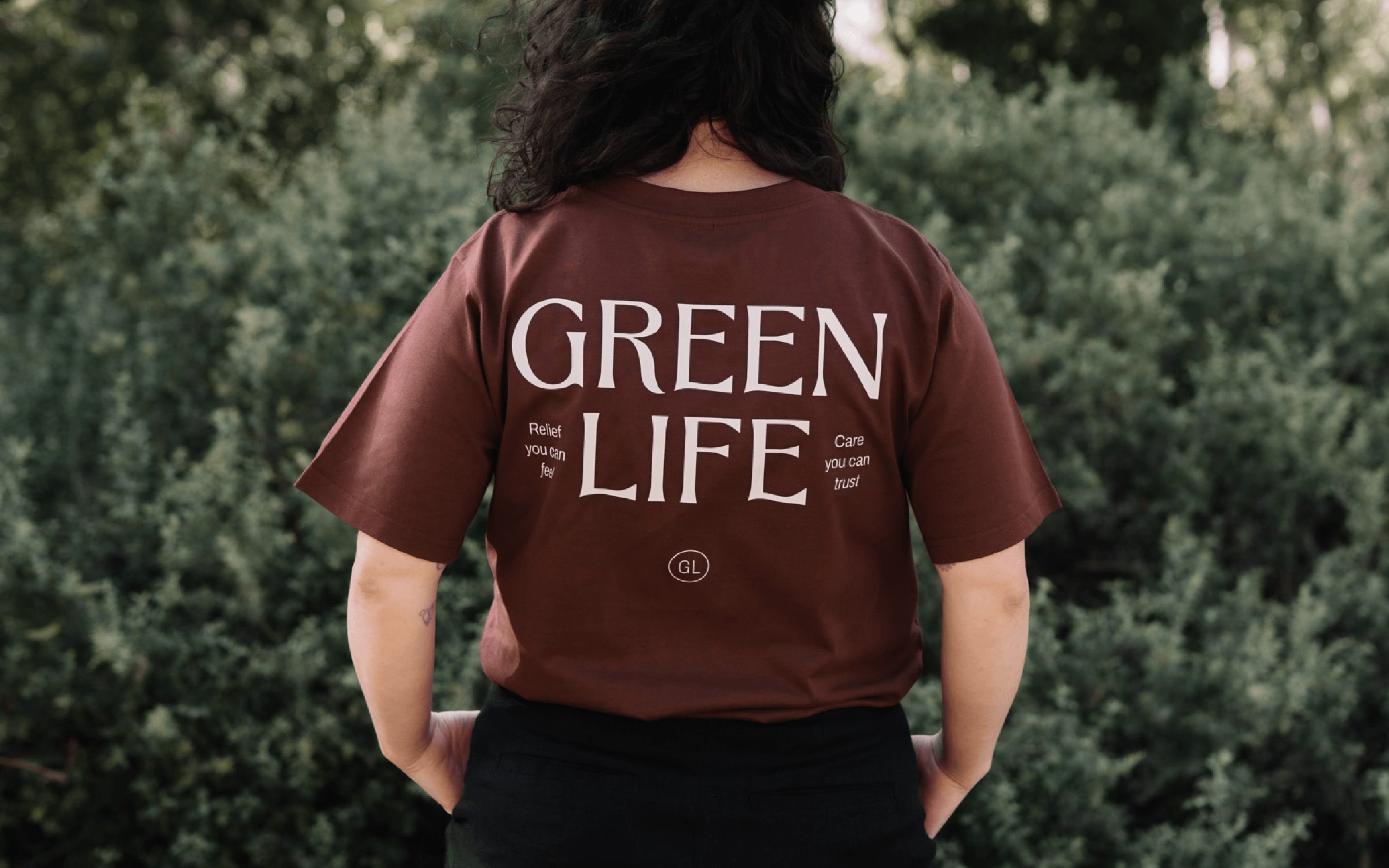 Branded t-shirt for an aged care massage therapy service, photographed on a person from behind, featuring the business name in large serif typography with tagline text and a monogram brand icon.