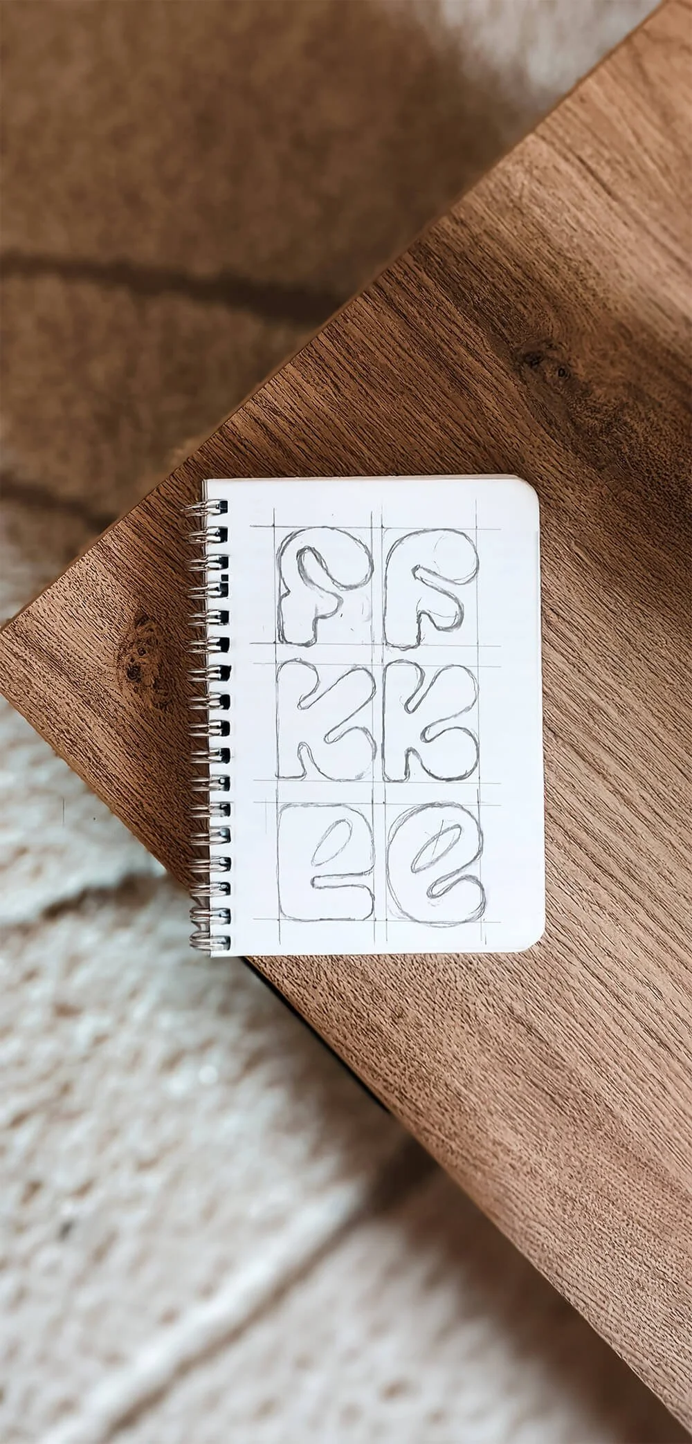 Sketches of custom typography for a fashion brand, featuring rounded bold letterforms drawn in a grid on a spiral notebook, styled on a timber table with a neutral textured background.