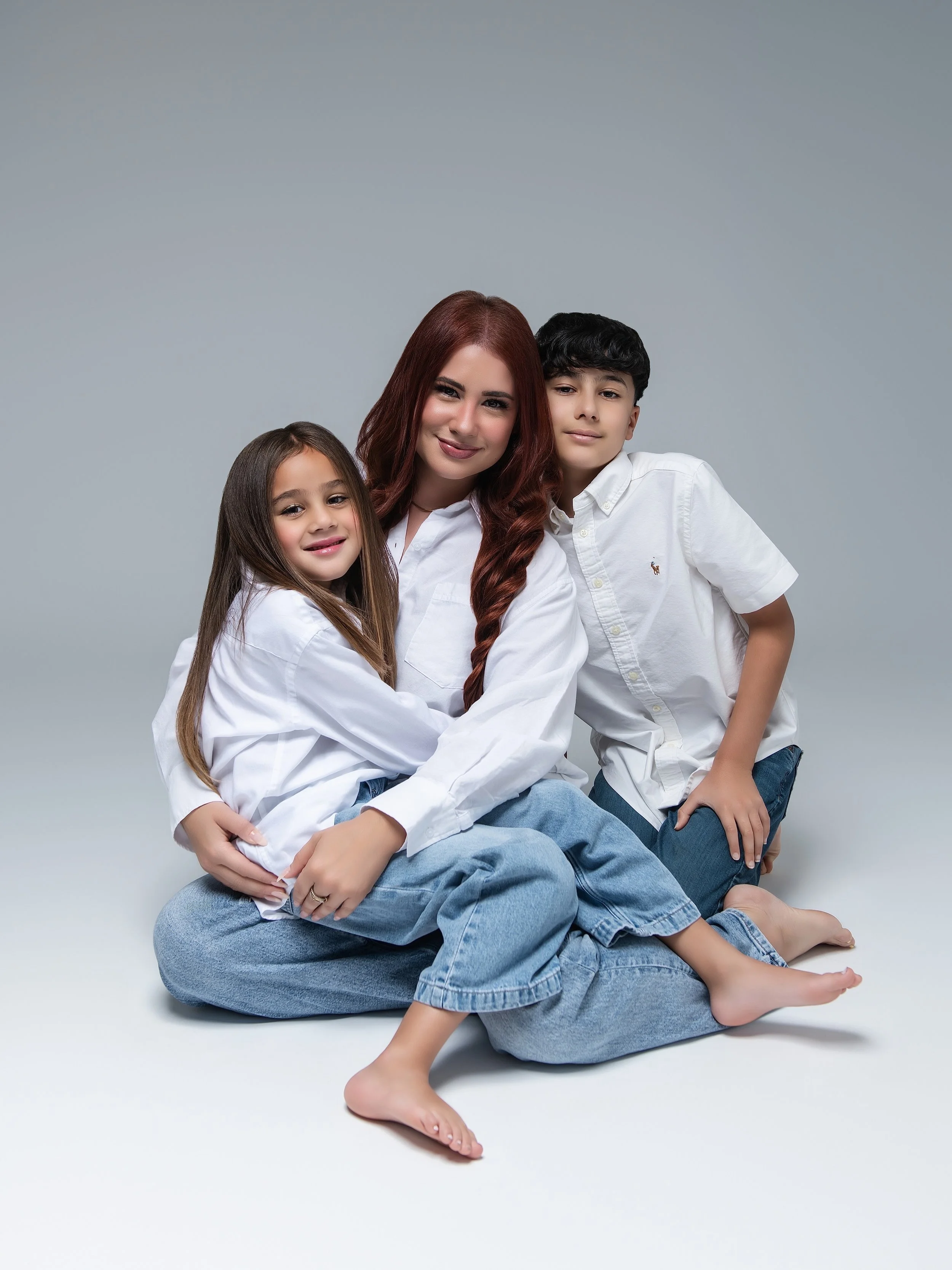 A woman with red hair and two children, a girl and a boy, sitting and hugging in a studio with a gray background, wearing white shirts and blue jeans.