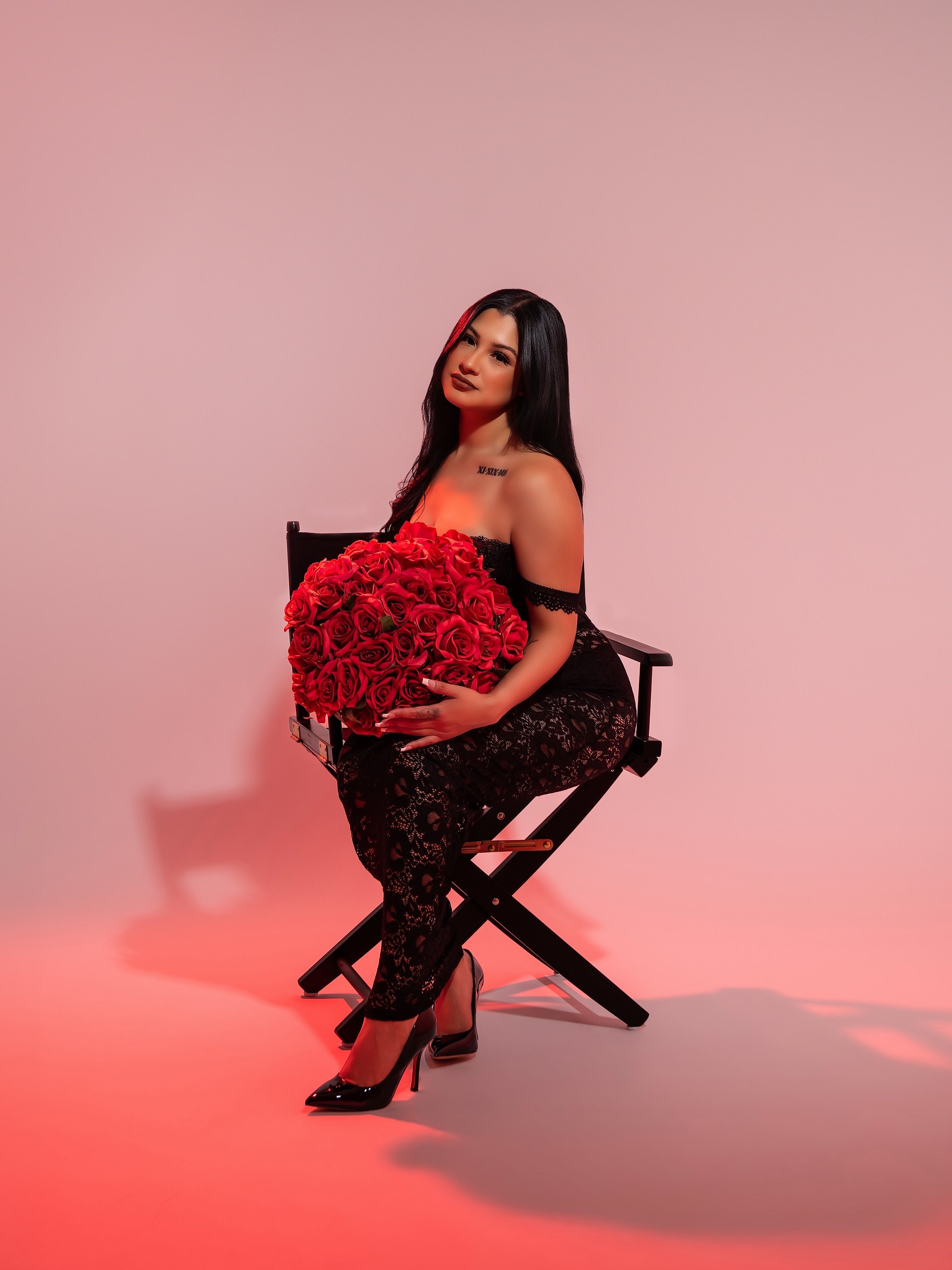 A woman with fair skin and long black hair sitting on a black director's chair, holding a large bouquet of red roses, wearing a black off-shoulder lace dress and black high heels, against a pink background with a red lighting effect.