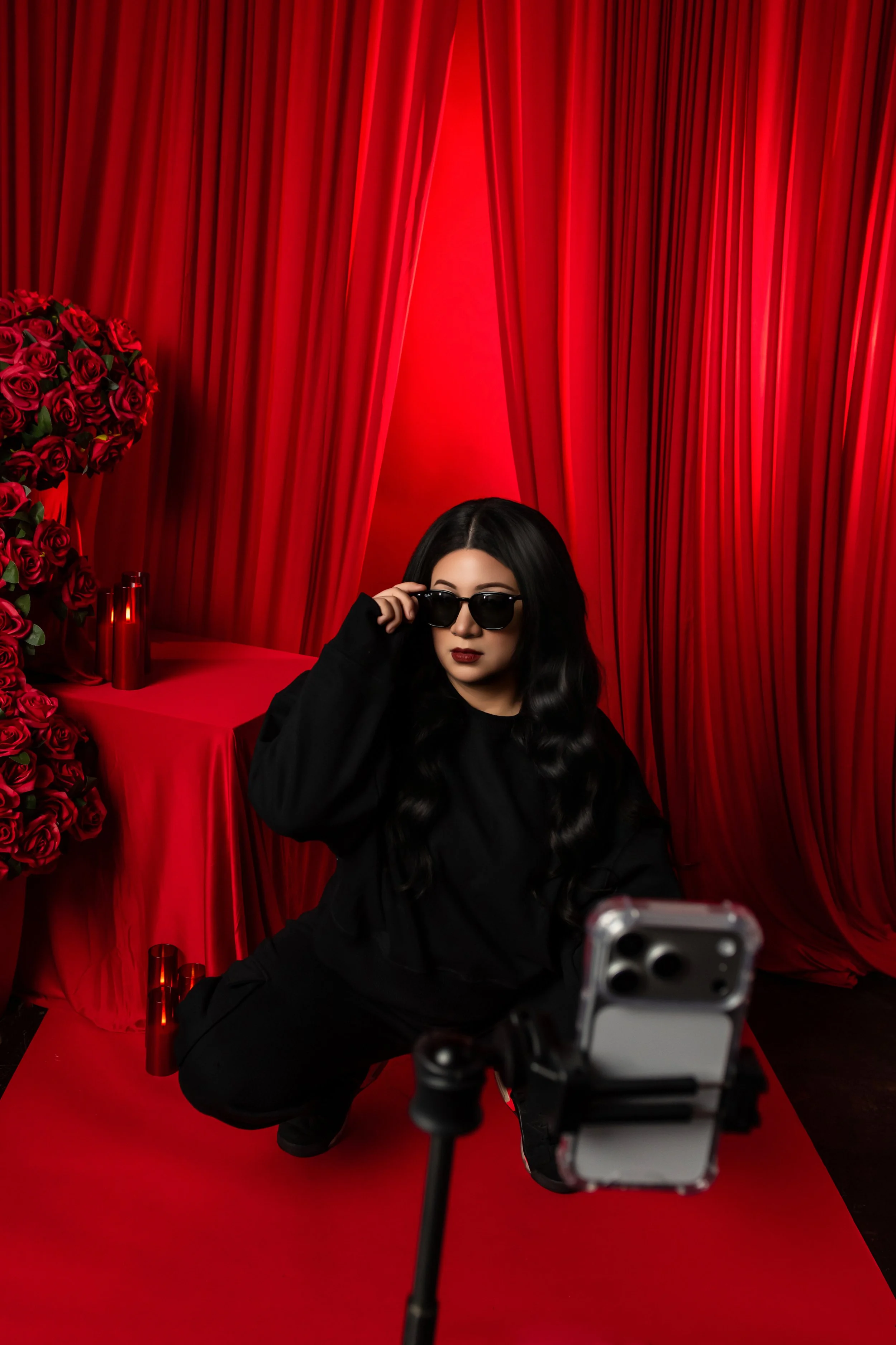 A woman with long black hair, wearing black sunglasses and a black outfit, sitting against a red curtain backdrop, taking a selfie with a smartphone on a tripod.