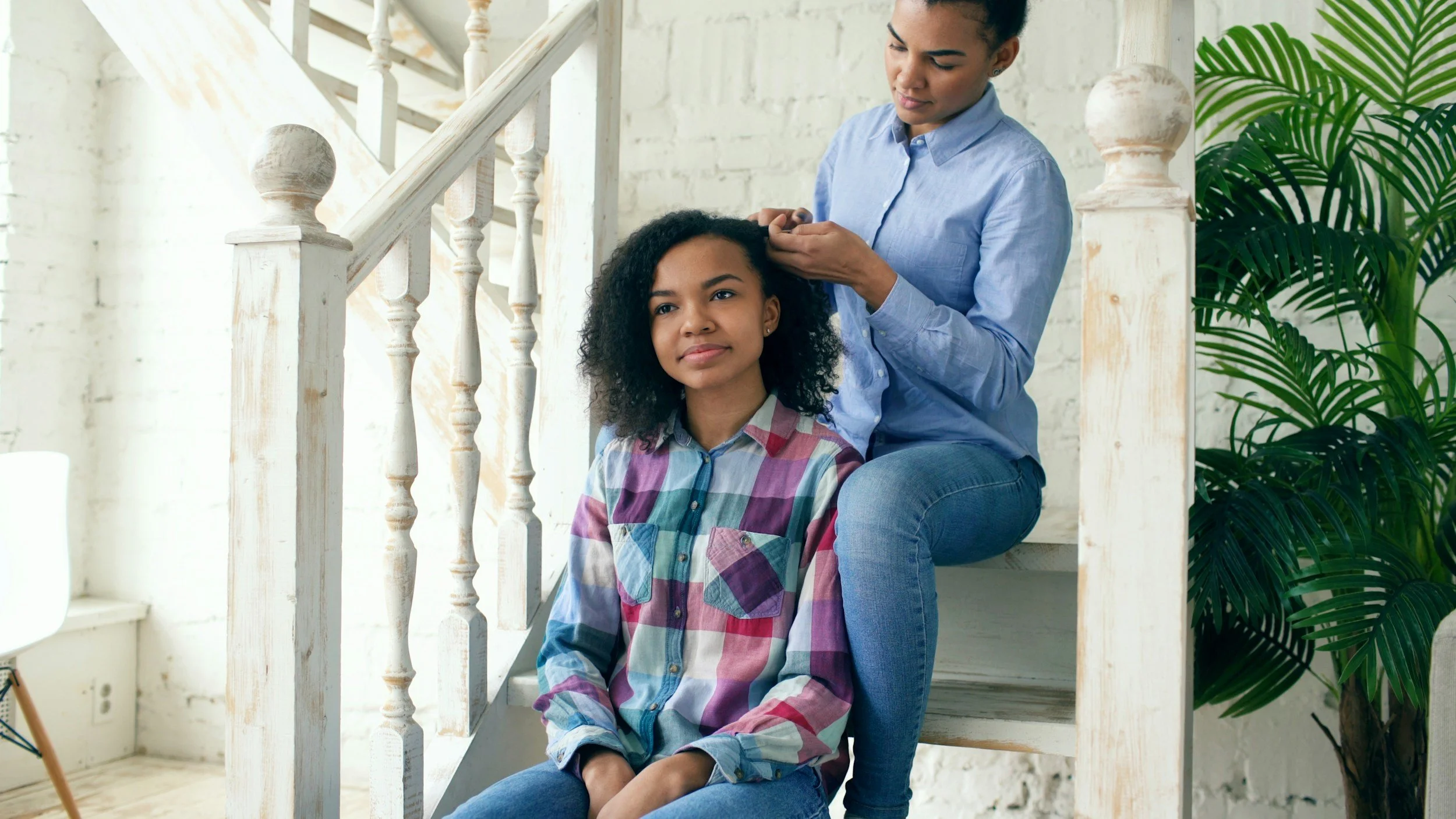 A woman styling a young girl's hair on a staircase in a bright, modern room with white brick walls and a large green plant nearby.