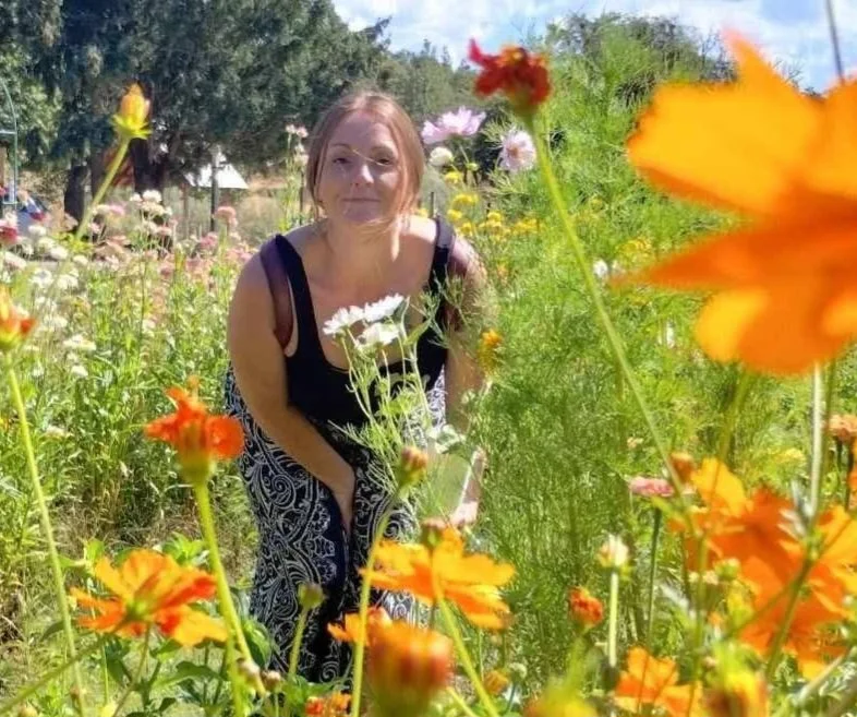 A woman with light skin and reddish-brown hair in a loose ponytail, wearing a black sleeveless top and patterned black and white pants, stands among colorful flowers in a garden on a sunny day. She is slightly bending forward, holding a flower, with a background of green bushes and trees.