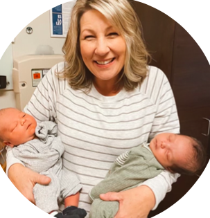 A woman smiling holds two newborn babies, one in each arm, in a room with medical equipment.