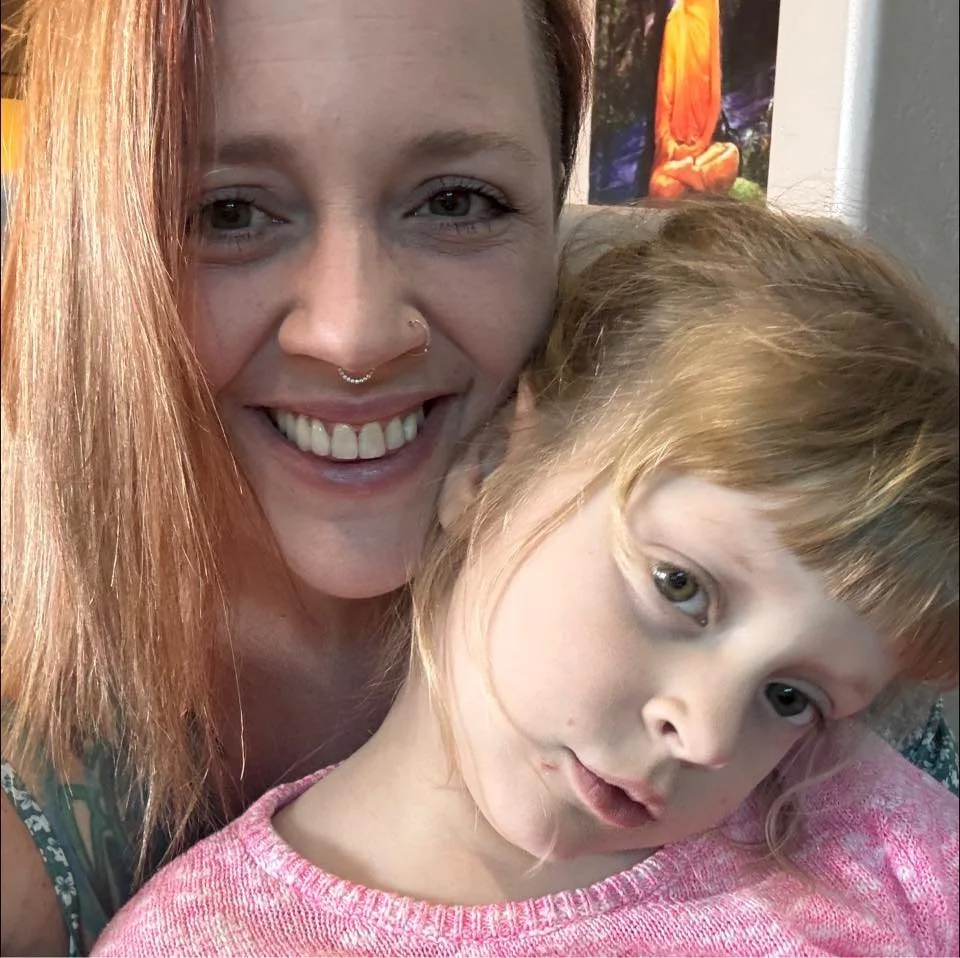 Two females, an adult woman with brown hair and a nose ring, and a young girl with blonde hair and green eyes, are close together taking a selfie. The woman is smiling and the girl looks slightly curious or concerned.