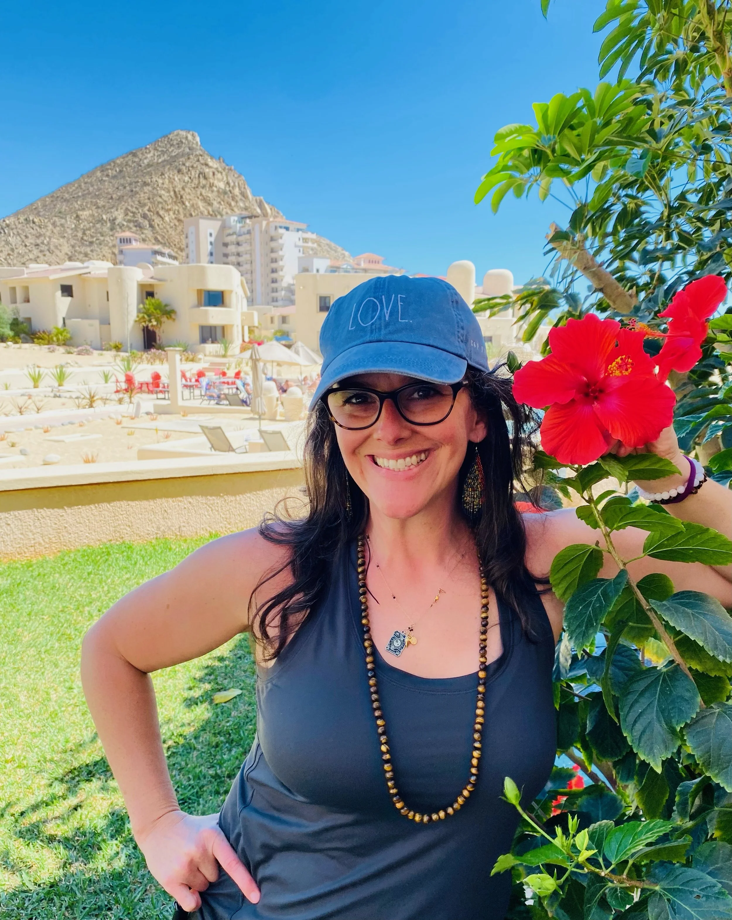A woman smiling and holding a red hibiscus flower outdoors, wearing glasses, a blue hat with 'LOVE' written on it, a black tank top, and jewelry, with a background of buildings, a mountain, and a garden.