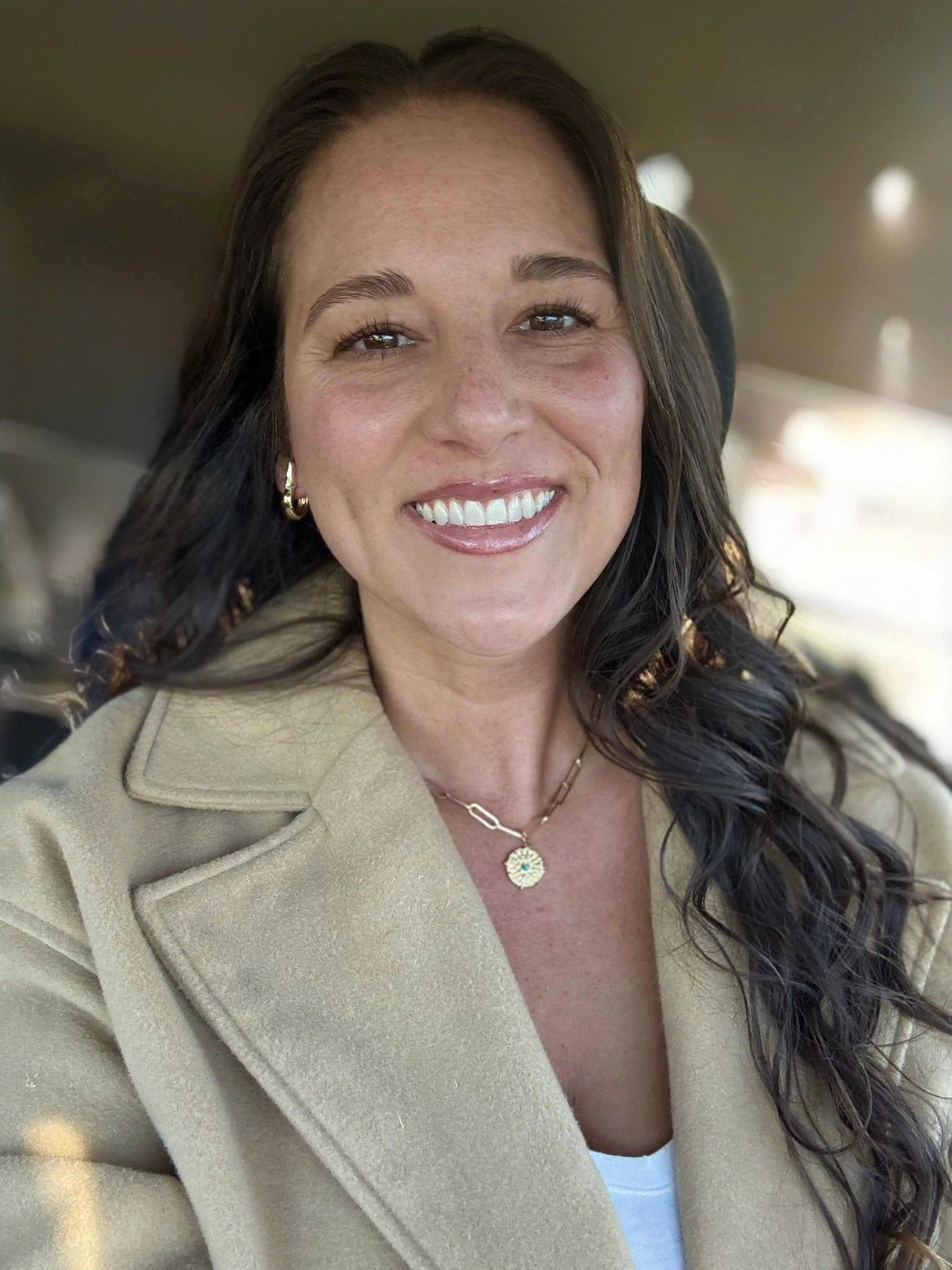A woman with long dark hair smiling inside a vehicle, wearing a beige coat, gold jewelry, and a white top.