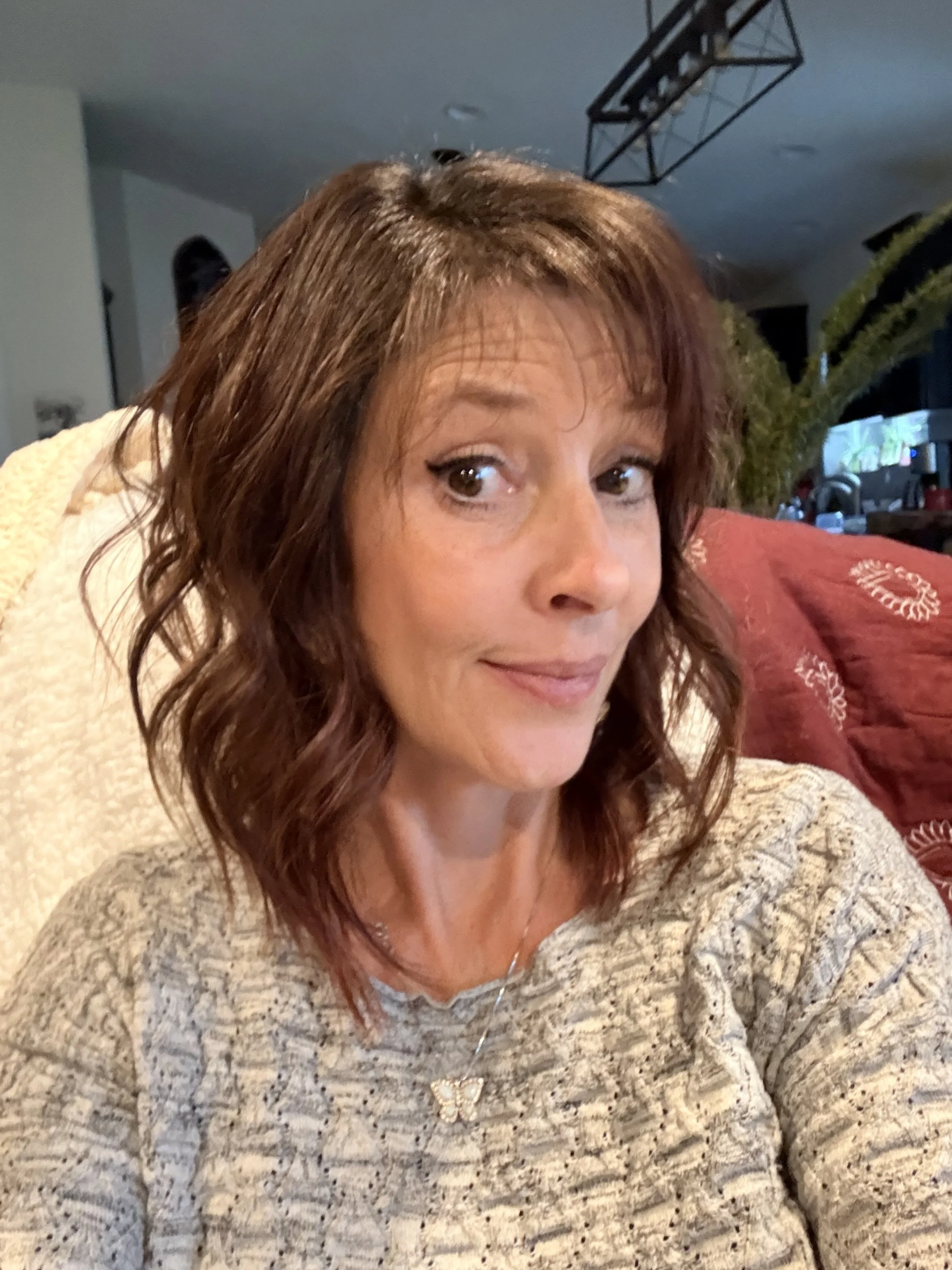 A woman with shoulder-length brown hair with reddish highlights taking a selfie in a cozy living room. She is wearing a beige sweater with a butterfly necklace, and behind her is a red patterned pillow, a plant, and part of a kitchen area. The lighting is warm and inviting.