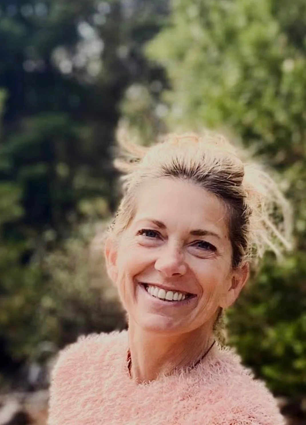 A woman smiling outdoors with greenery in the background.