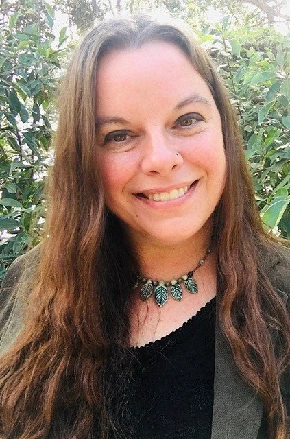 A woman with long brown hair, smiling, wearing a black top, a necklace, and a jacket, standing outdoors with greenery in the background.