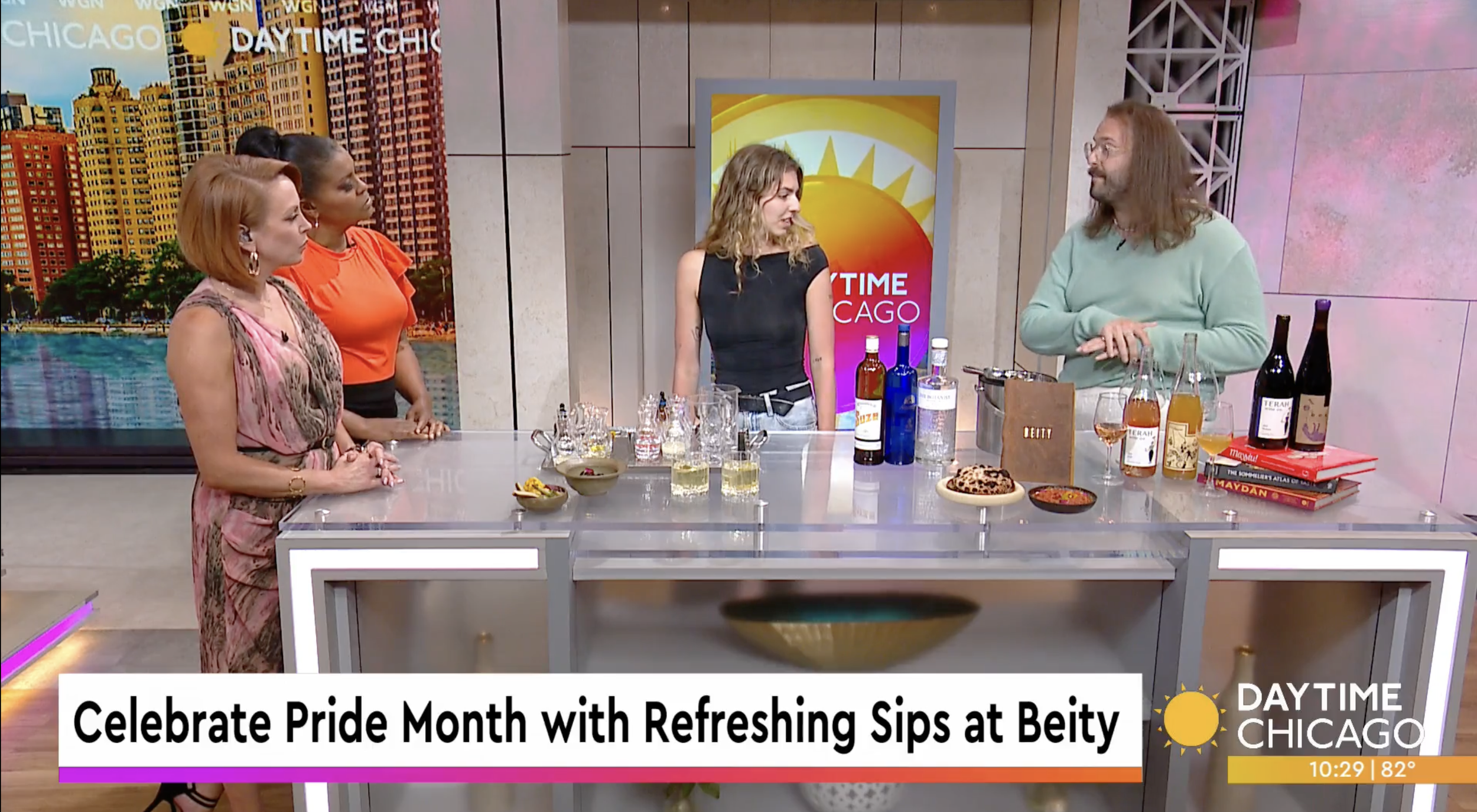 A daytime television segment on a news show features four women standing around a table with various drinks and snacks, discussing Pride Month with a guest, Beity. The set has cityscape images and a colorful background with the show’s logo, "Daytime Chicago."