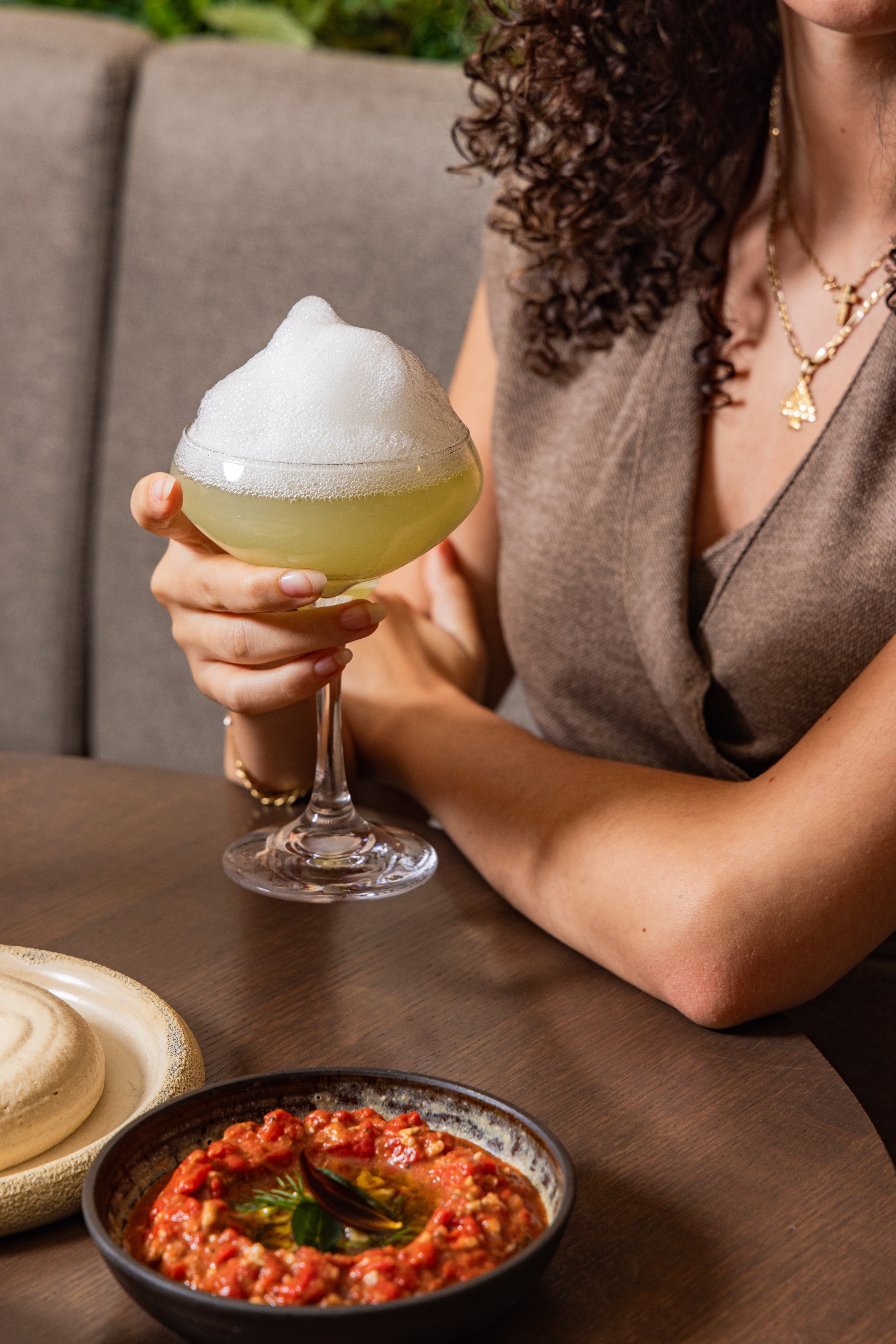 A woman with curly hair and gold necklaces holds a glass of greenish cocktail topped with foamy bubbles, sitting at a table with various dishes including a bowl of tomato-based dip, bread rolls, and other food items.
