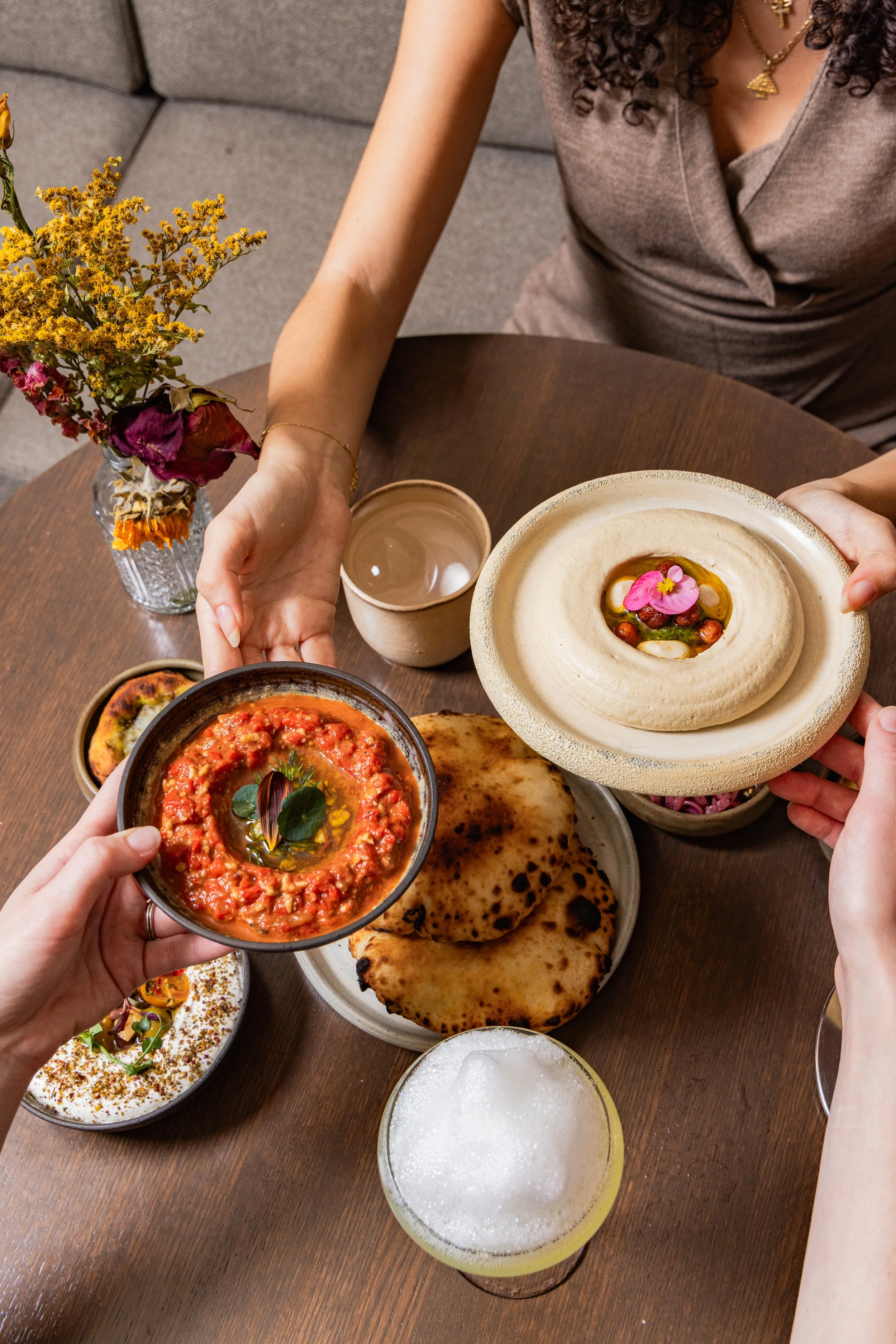 A serving of spicy tomato-based dip with herbs in a small bowl, two naan breads, several other bowls with Indian dishes, and three drinks on a wooden table, with a person reaching for the food.