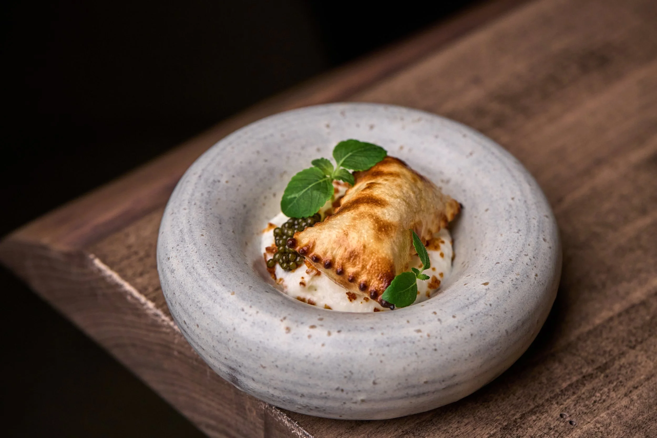 A savory tart with a golden, crispy crust, topped with caviar, fresh green leaves, and a dollop of cream, served on a white speckled ceramic plate.