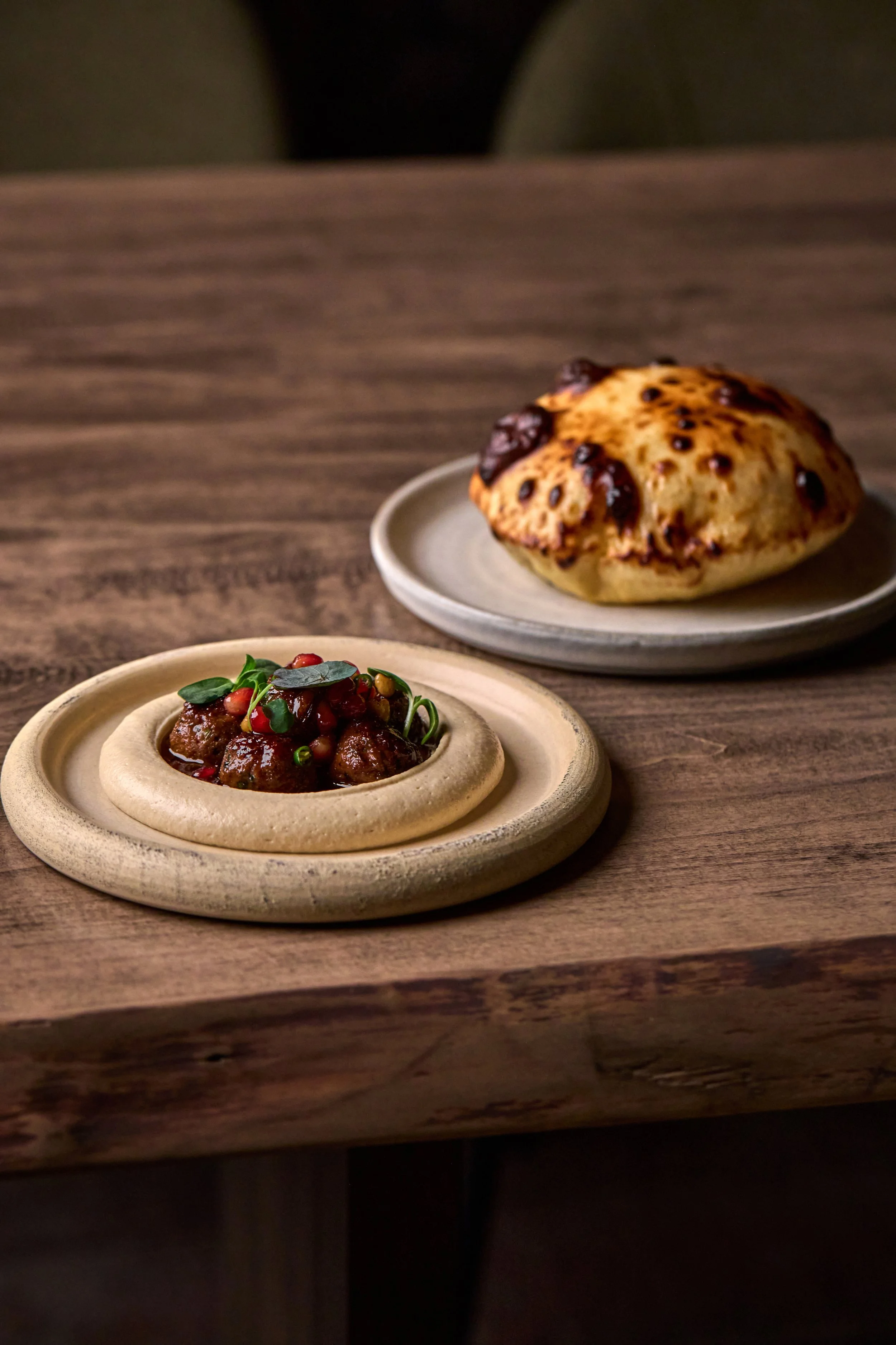 A baked chocolate chip scone on a gray plate and a bowl of meatball stew garnished with greens and pomegranate seeds on a wooden table.