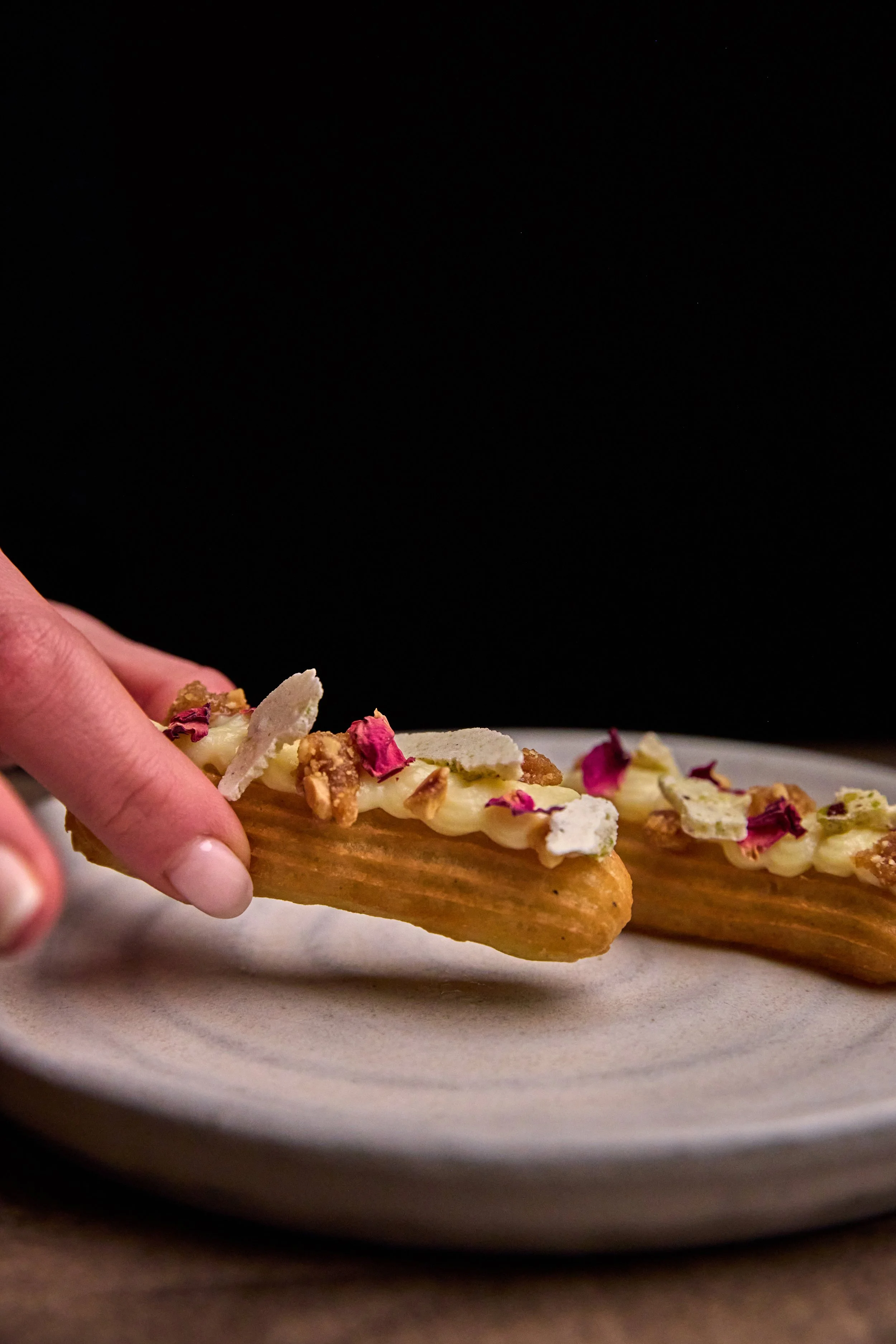 Close-up of a person holding a piece of churro topped with white frosting, chopped nuts, and rose petals on a white plate against a black background.