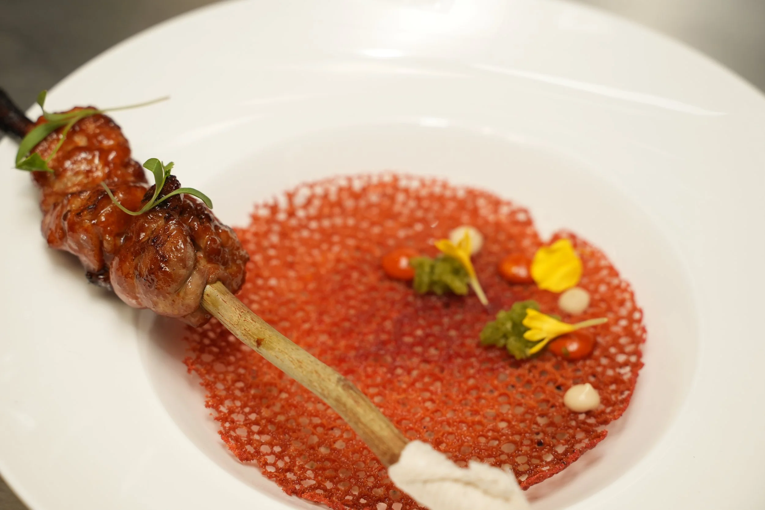 Close-up of a gourmet figwood skewer with glazed sweetbread resting on a vibrant red tuille, garnished with shattah, Aleppo and lemon tahini
