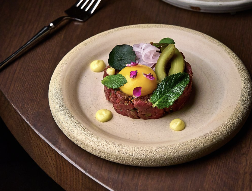 A plate of beef tartare topped with a raw egg yolk, garnished with green herbs and edible flower petals, served with small dollops of yellow sauce, on a beige ceramic plate with a fork nearby.