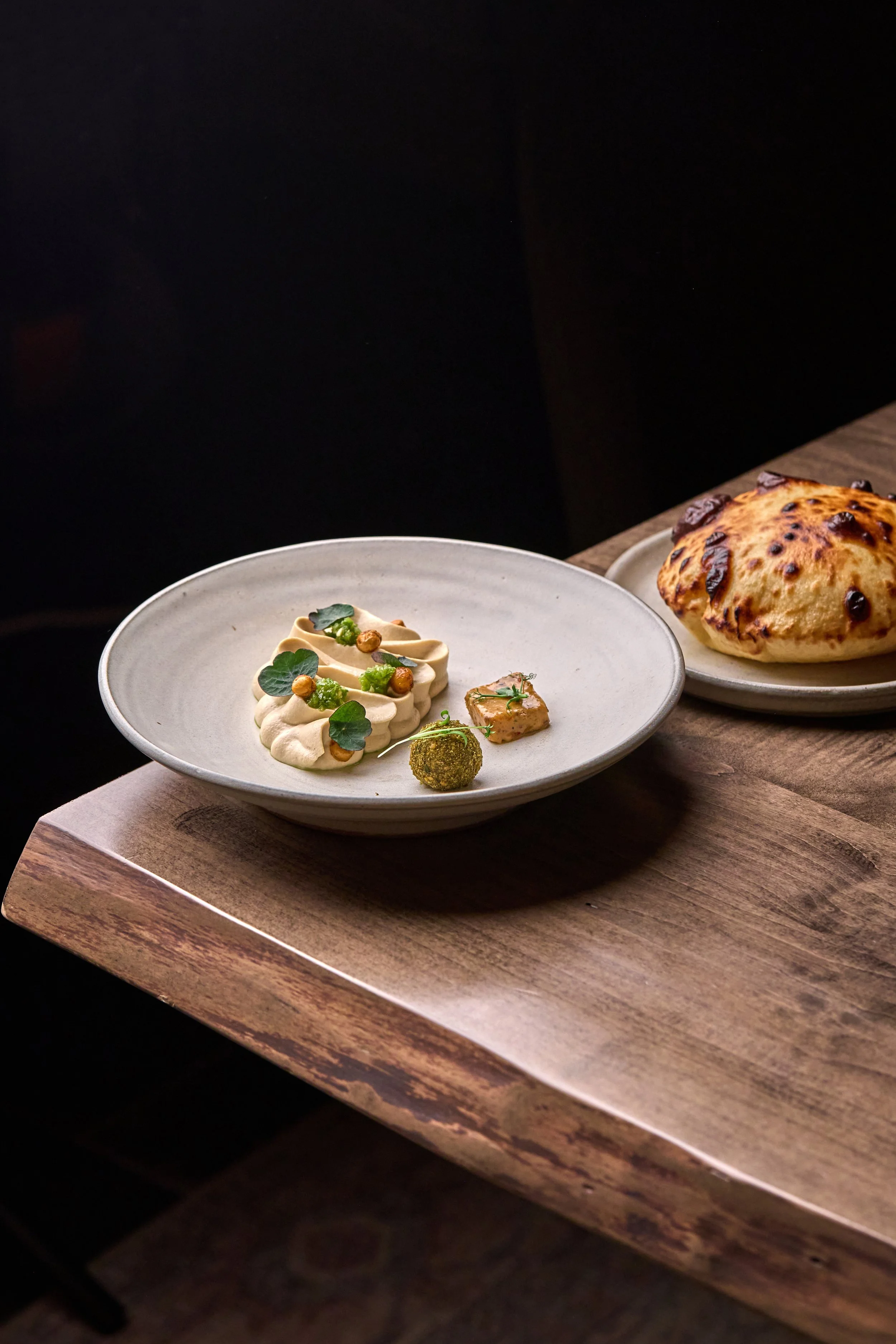 Close-up of a fine dining presentation with a plate of creamy appetizer garnished with microgreens and small edible flowers, alongside a toasted bread roll with dark spots, placed on a wooden serving board.