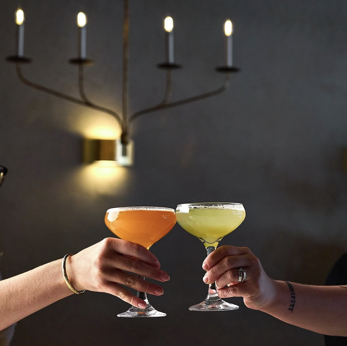 Two people toasting with cocktails in coupe glasses, one with orange drink and the other with pale green drink, against a dark background with a modern chandelier.