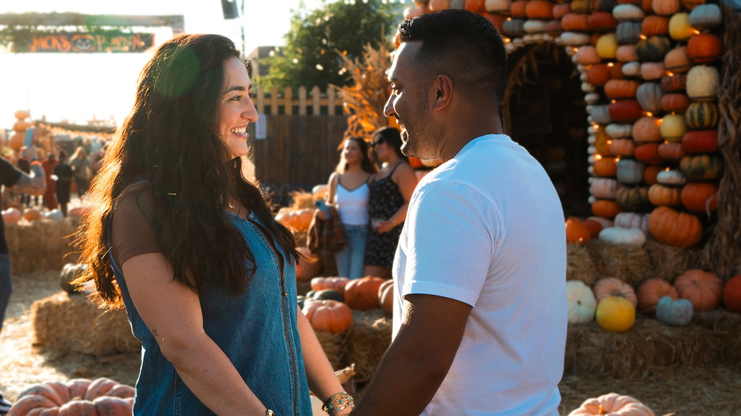 A couple is smiling at each other at an outdoor pumpkin patch, holding hands. There are pumpkins and hay bales in the background with other people visiting the pumpkin patch on a sunny day.