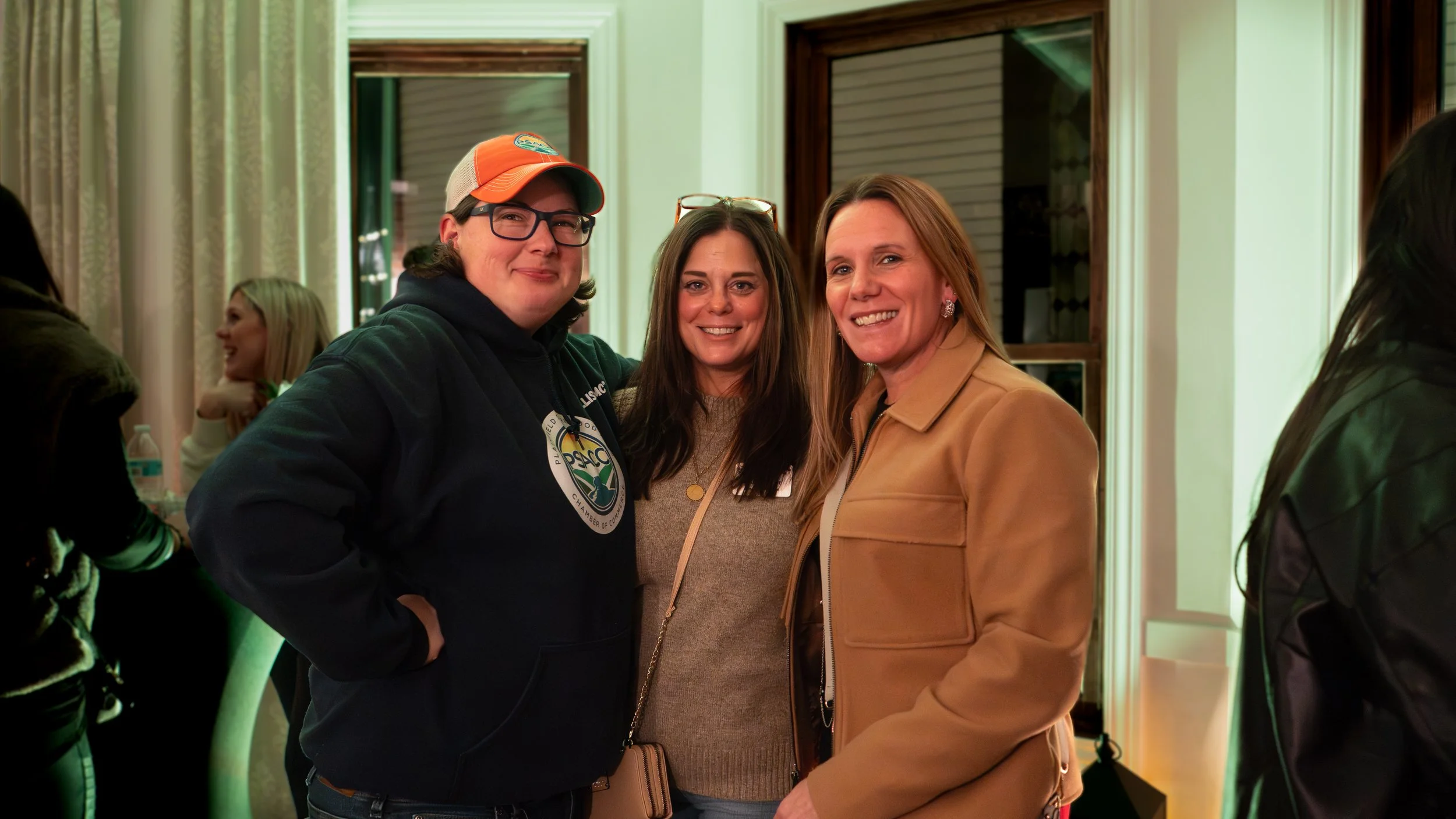Three women smiling at an indoor event, with people in the background, one wearing a hoodie and cap, another in a tan jacket, and the third in a beige sweater, standing near windows with curtains.