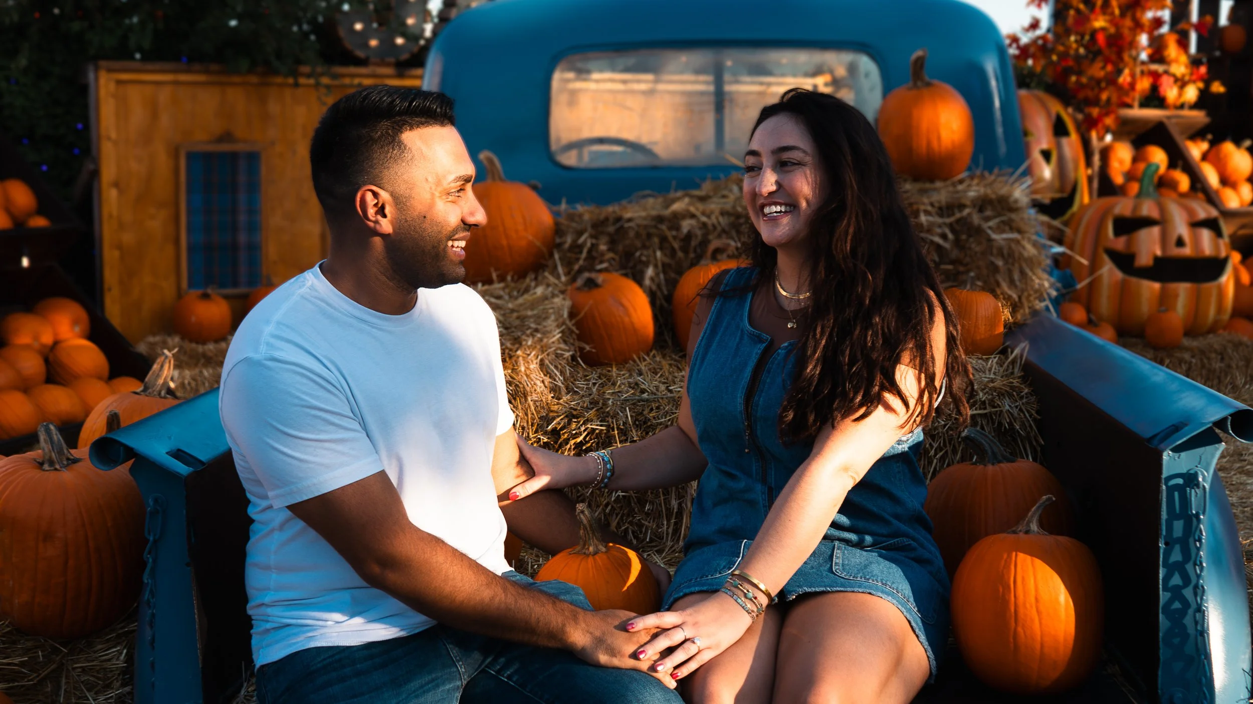 A couple sitting in the back of a small trailer filled with pumpkins and hay, smiling at each other during fall season.