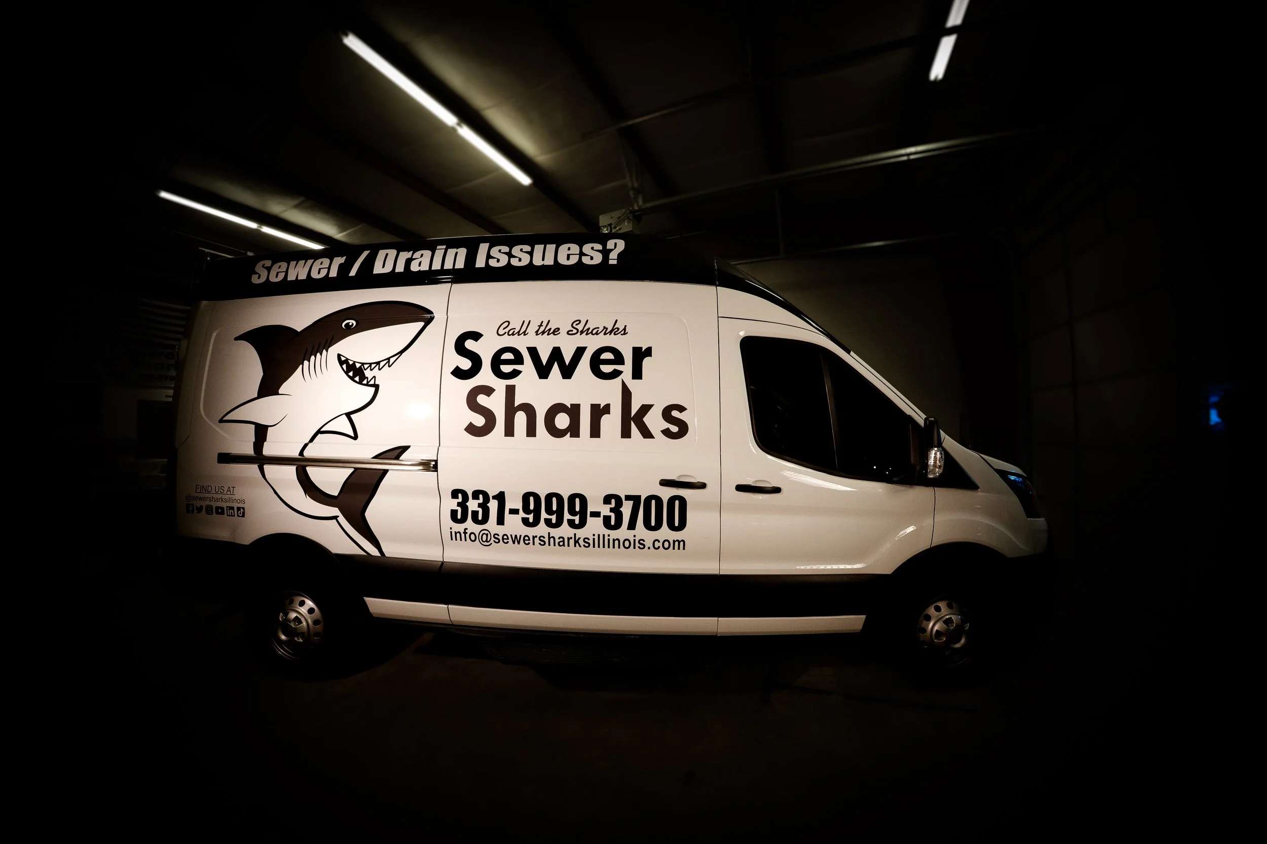 Sewer Sharks service van inside a dark garage, featuring the company's logo with a cartoon shark, contact number, and email address for sewer and drain issues.