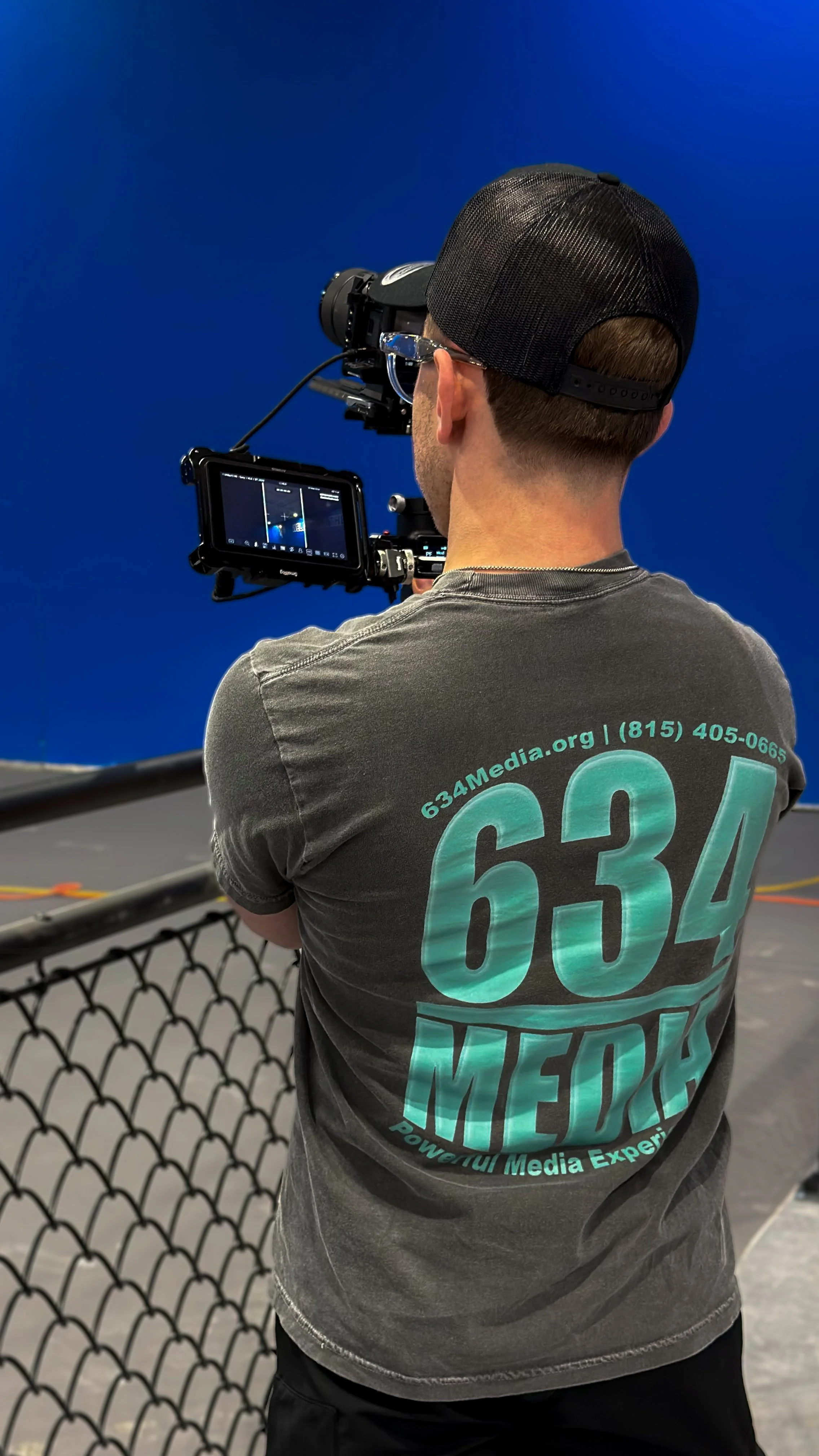 A person wearing a grey t-shirt and a black cap using a video camera, filming against a blue background.