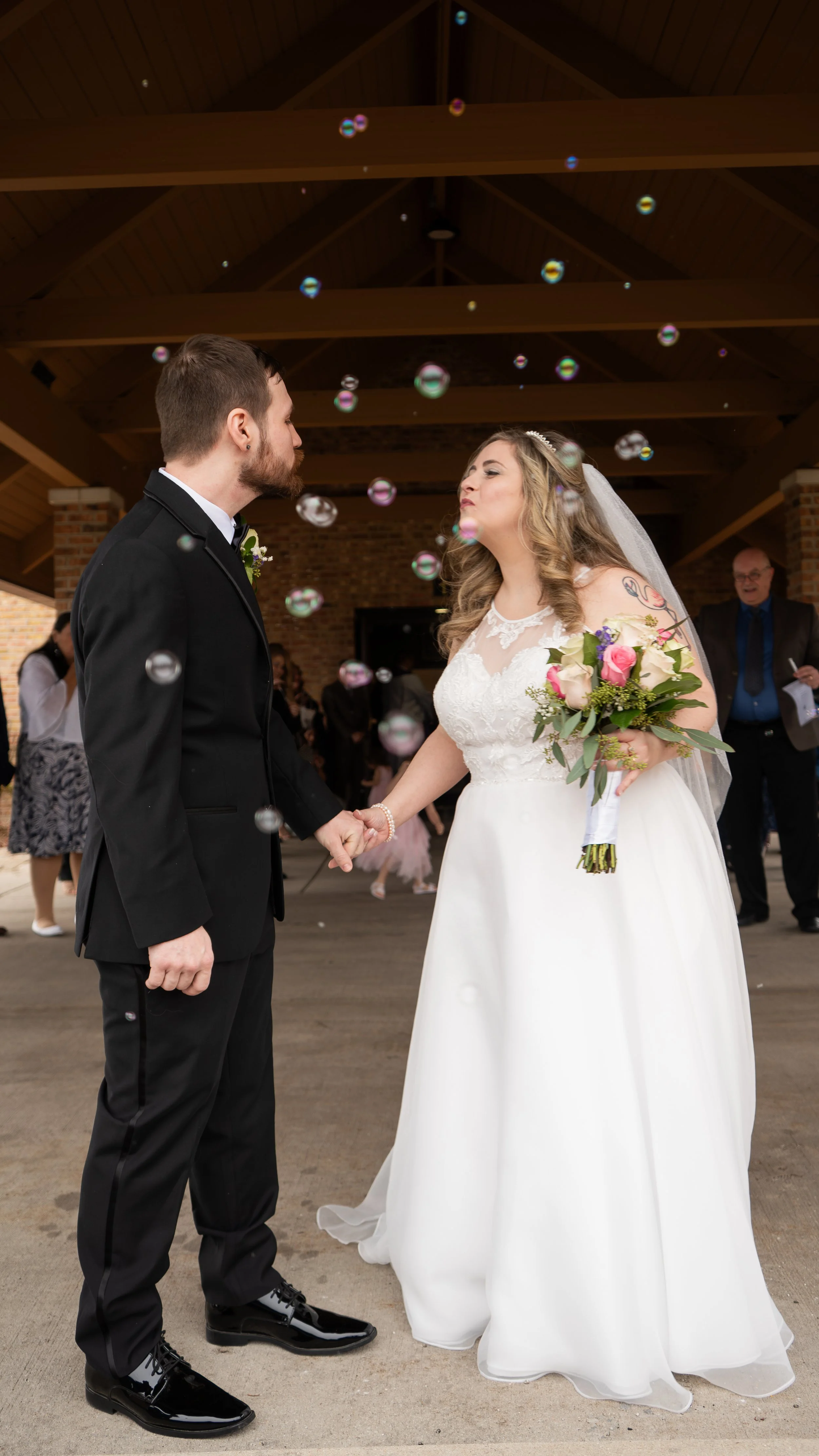 Bride and groom holding hands during wedding ceremony, surrounded by bubbles and wedding guests, under a wooden pavilion.