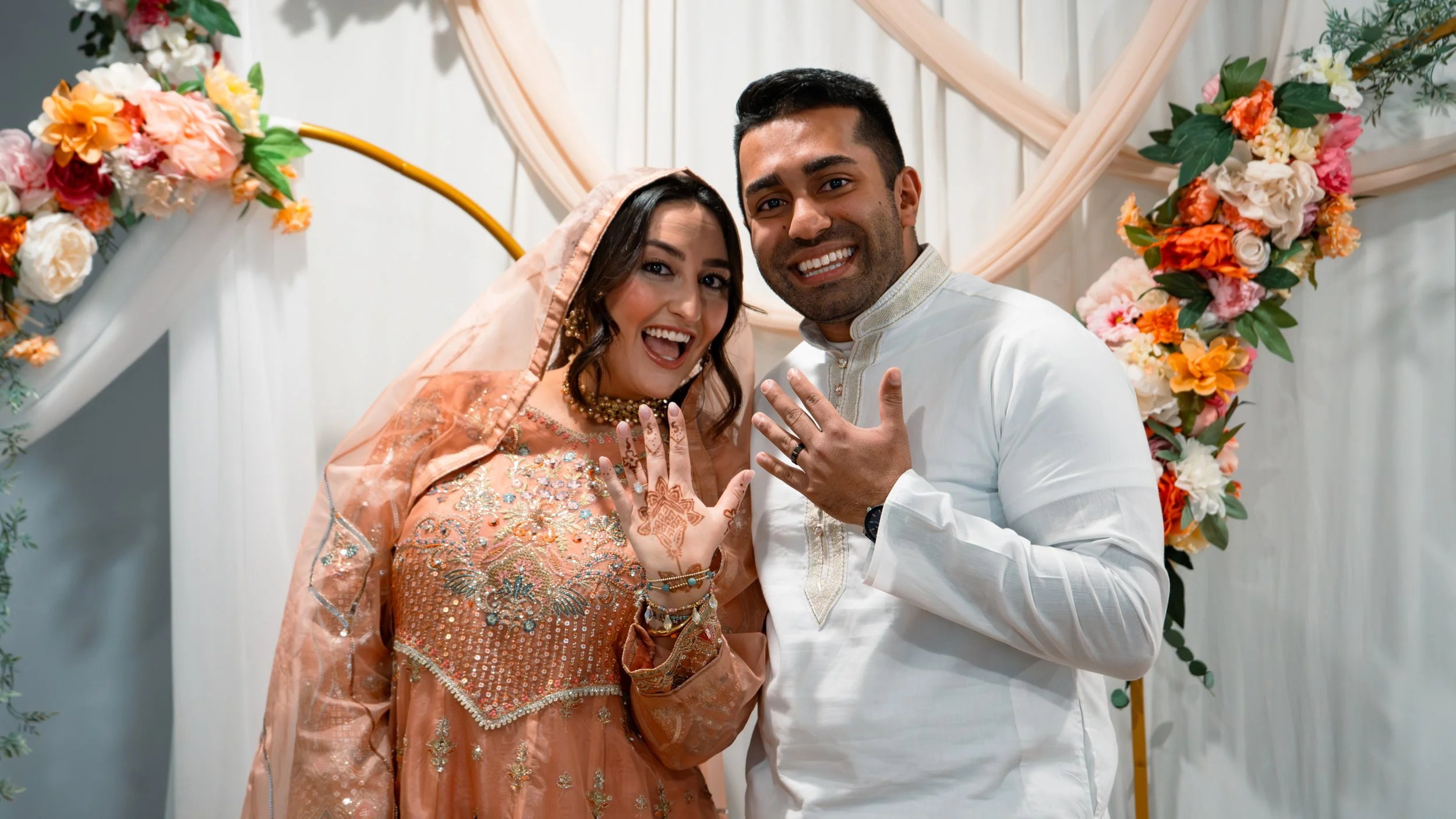 A happy couple showing their wedding rings at a wedding ceremony. The woman is dressed in a peach traditional outfit with embroidery, and the man in a white traditional suit. There are floral decorations in the background.