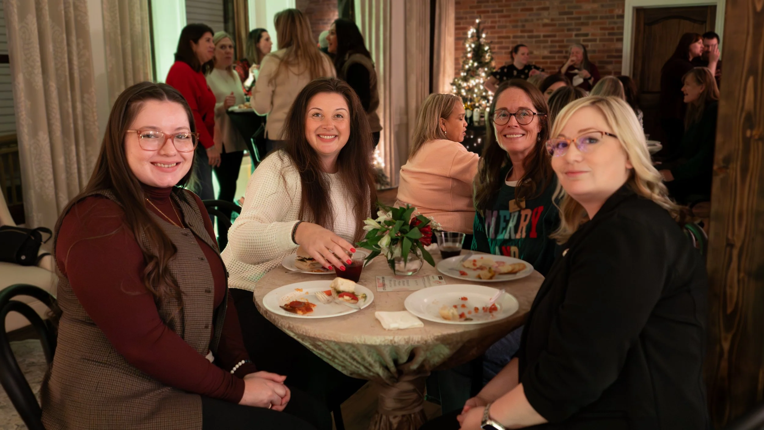 Four women sitting around a table at a holiday gathering, smiling, with a decorated Christmas tree and other people in the background.