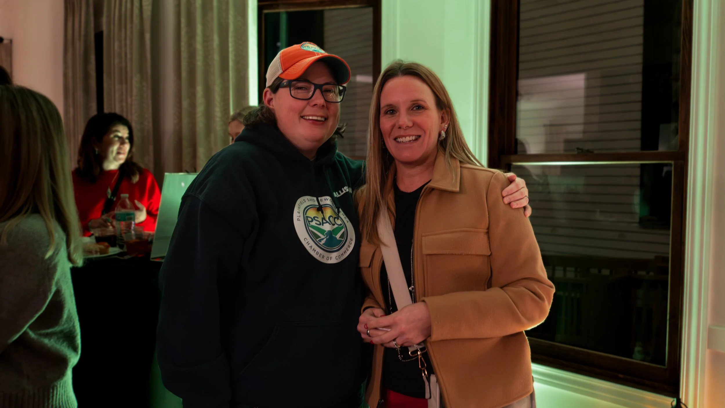 Two women smiling and posing together indoors, one wearing a hoodie with a logo and the other in a tan jacket, with other people and a table in the background.