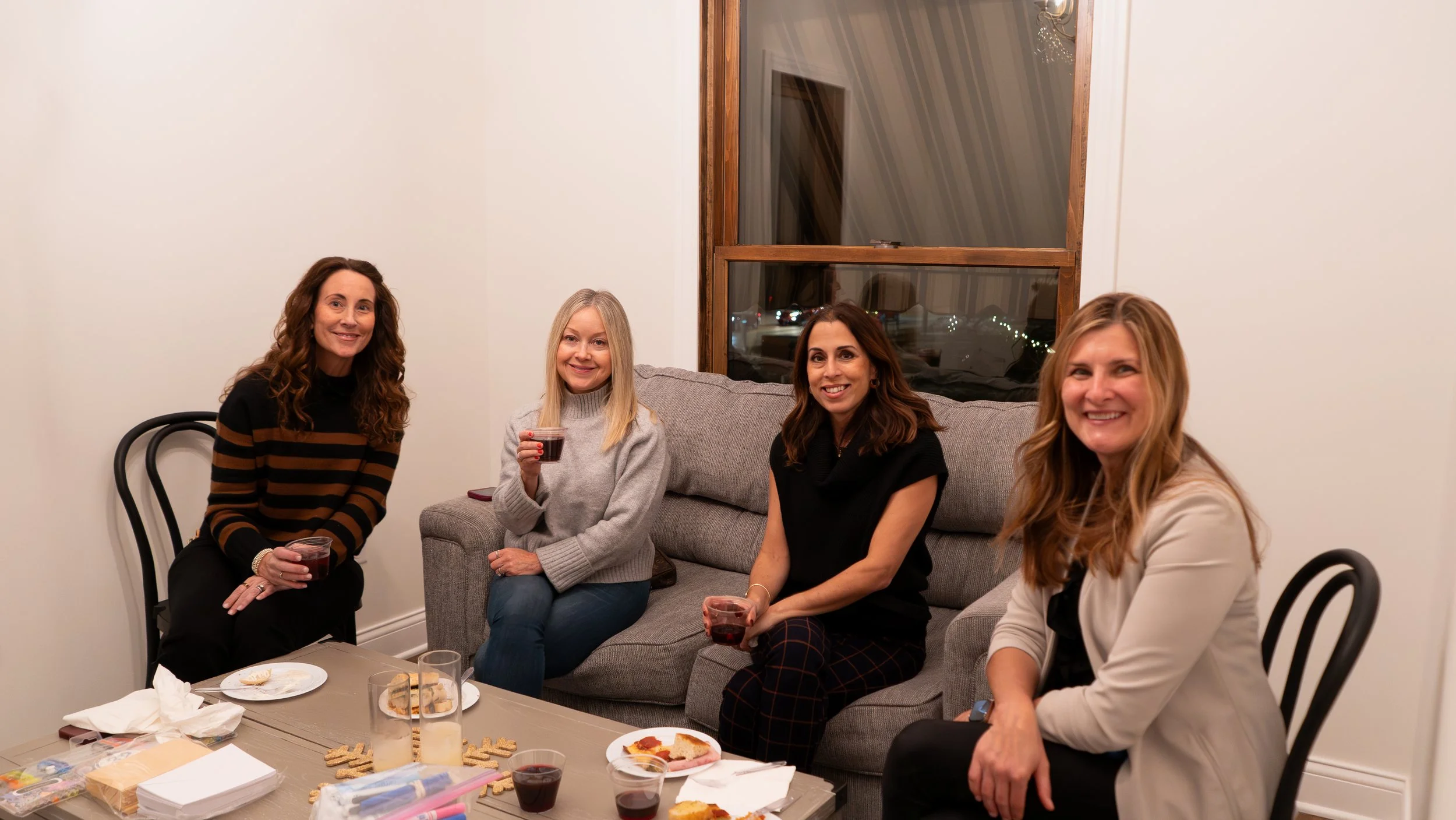 Four women sitting in a living room, smiling and holding drinks, with a table of snacks in front of them.