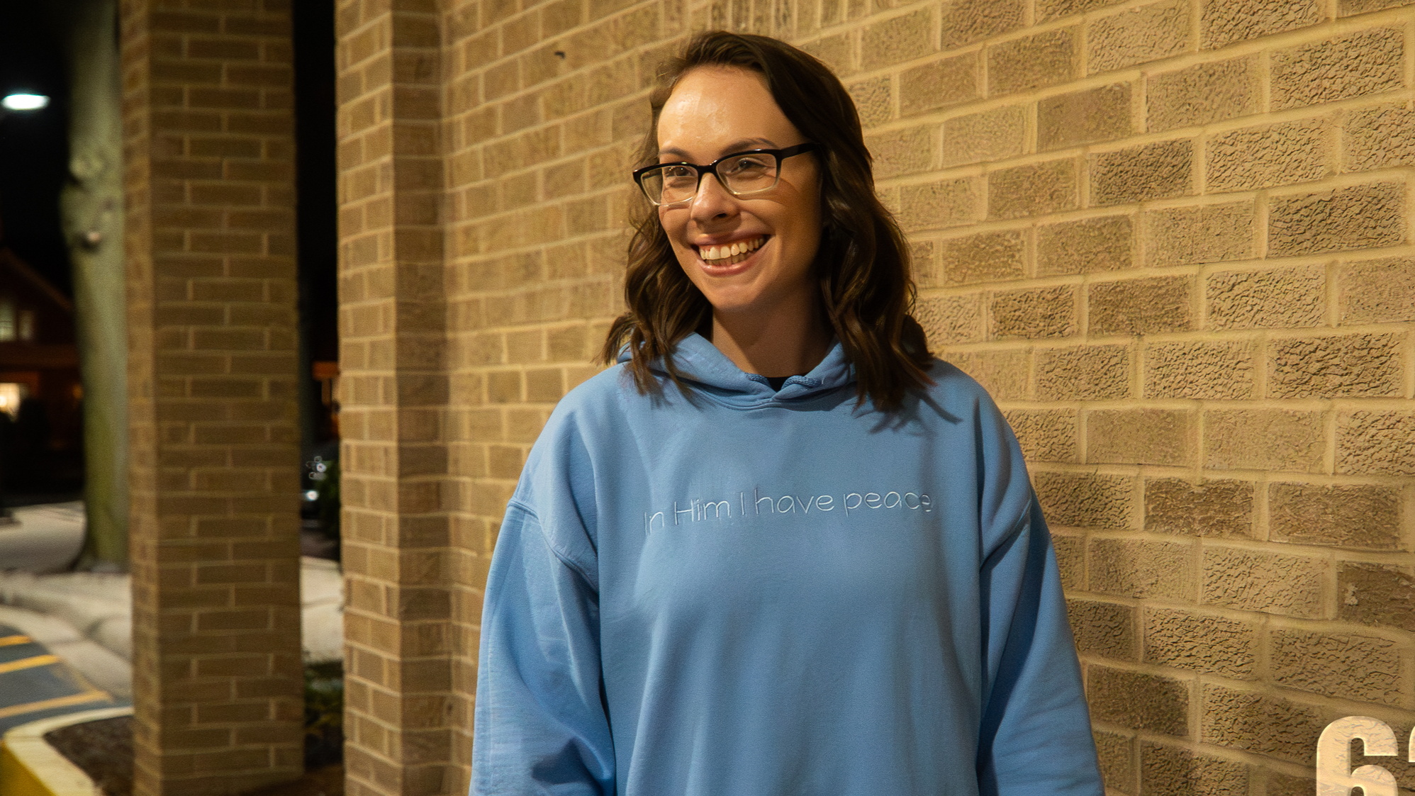 Young woman with glasses smiling, wearing a light blue hoodie with white text, standing outside against a brick wall at night.
