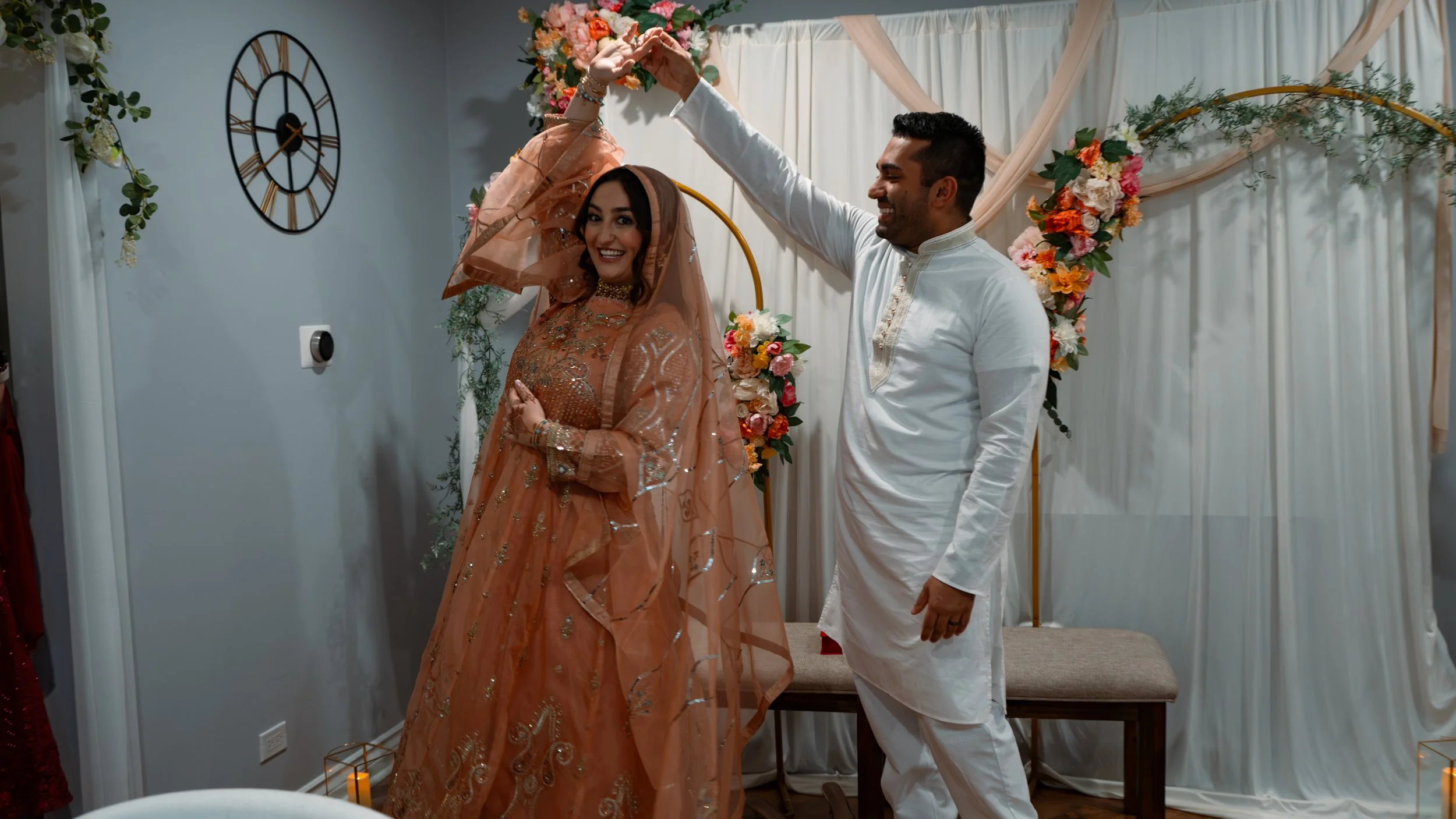 A couple dancing at a wedding, with the man twirling the woman under his hand. The woman is wearing a peach-colored traditional dress with embroidery and a veil. The man is in a white traditional outfit. They are indoors with floral decorations and a