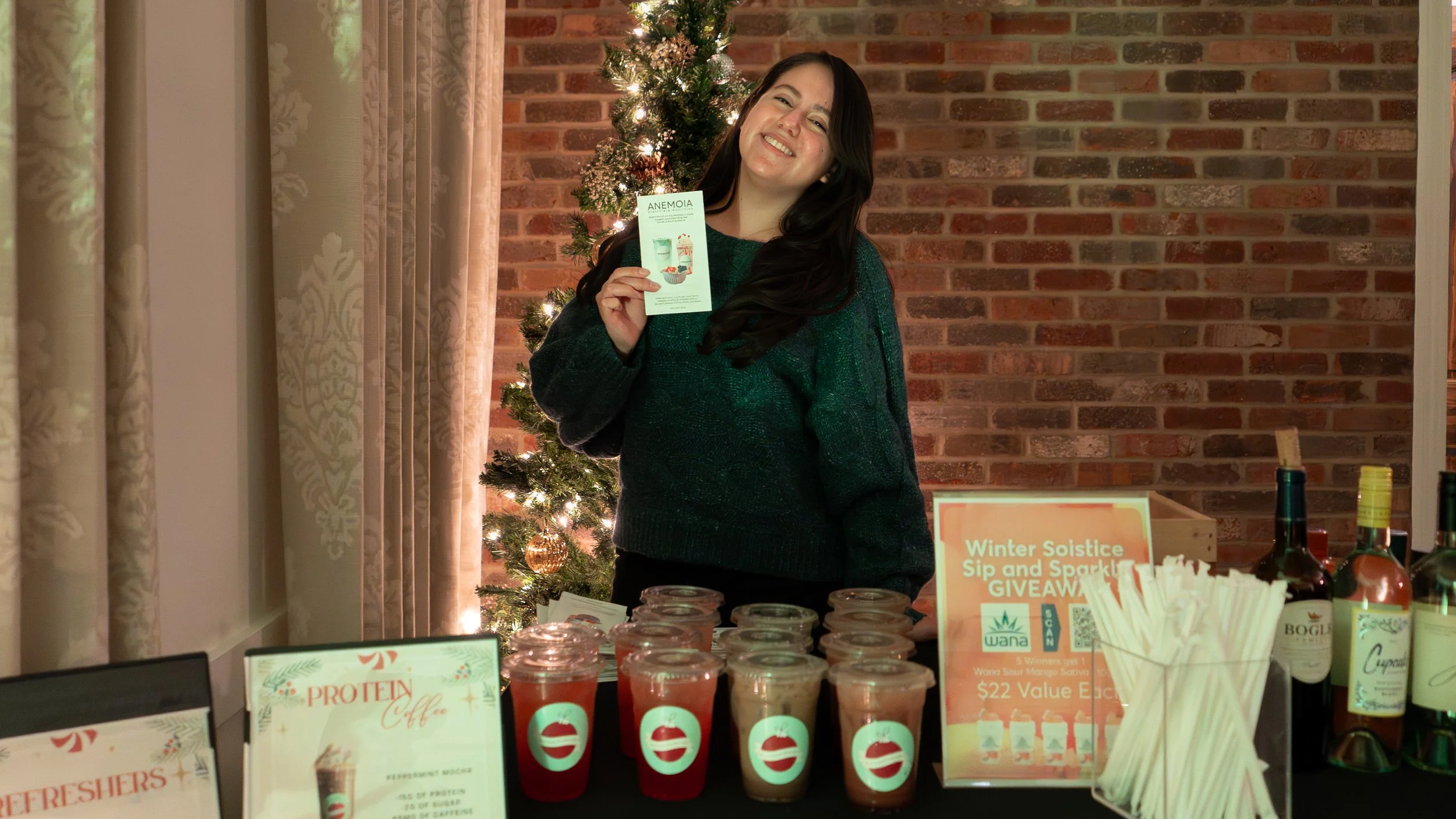 A woman smiling at a holiday event booth, holding a flyer, with a Christmas tree behind her decorated with lights and gold ornaments. The booth displays various drinks, including protein coffee and other beverages, with promotional signs for a winter