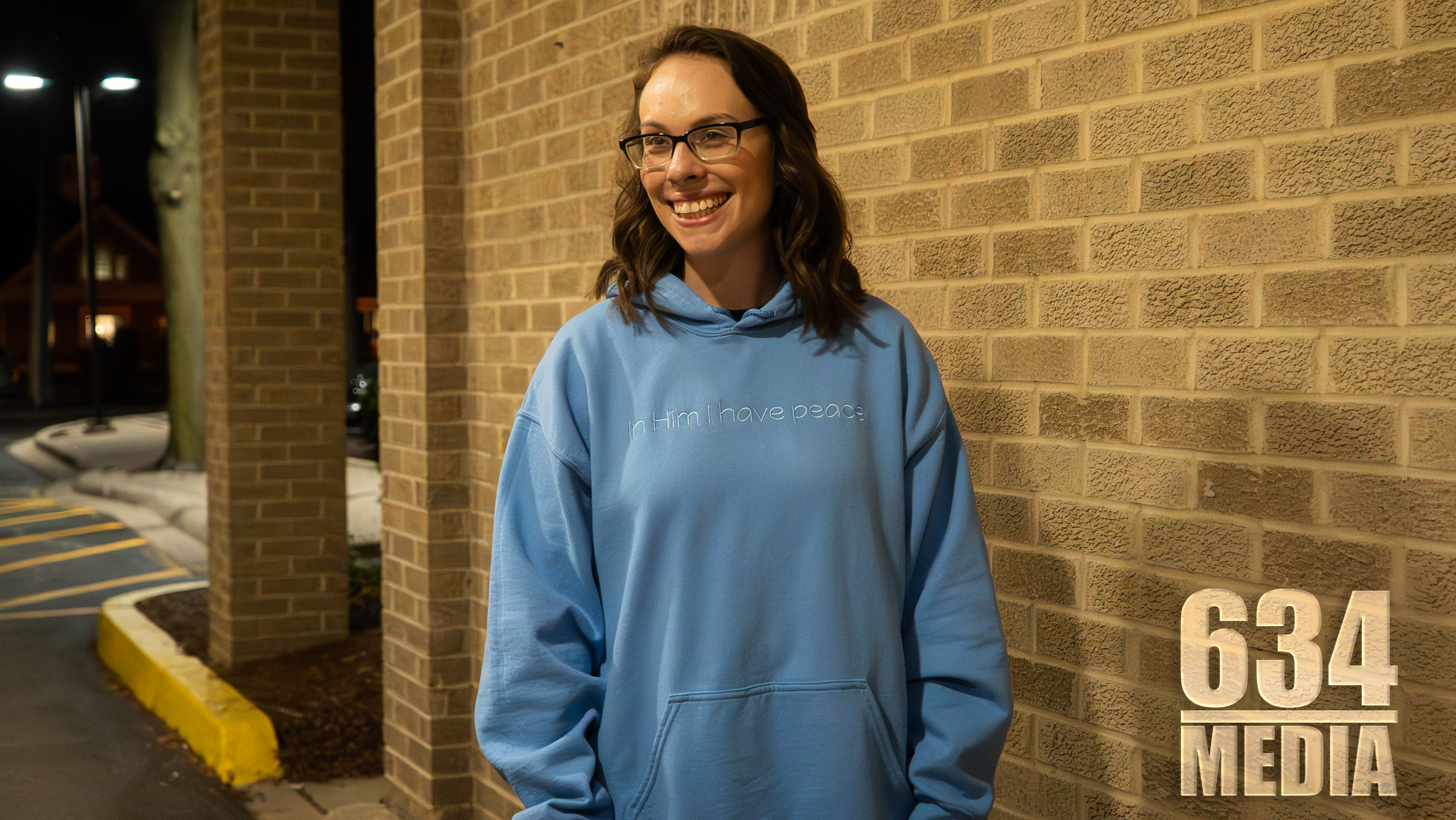 A young woman with glasses and brown wavy hair smiling while standing next to a brick wall at night. She is wearing a light blue hoodie with the embroidered text 'In Him I have peace.'
