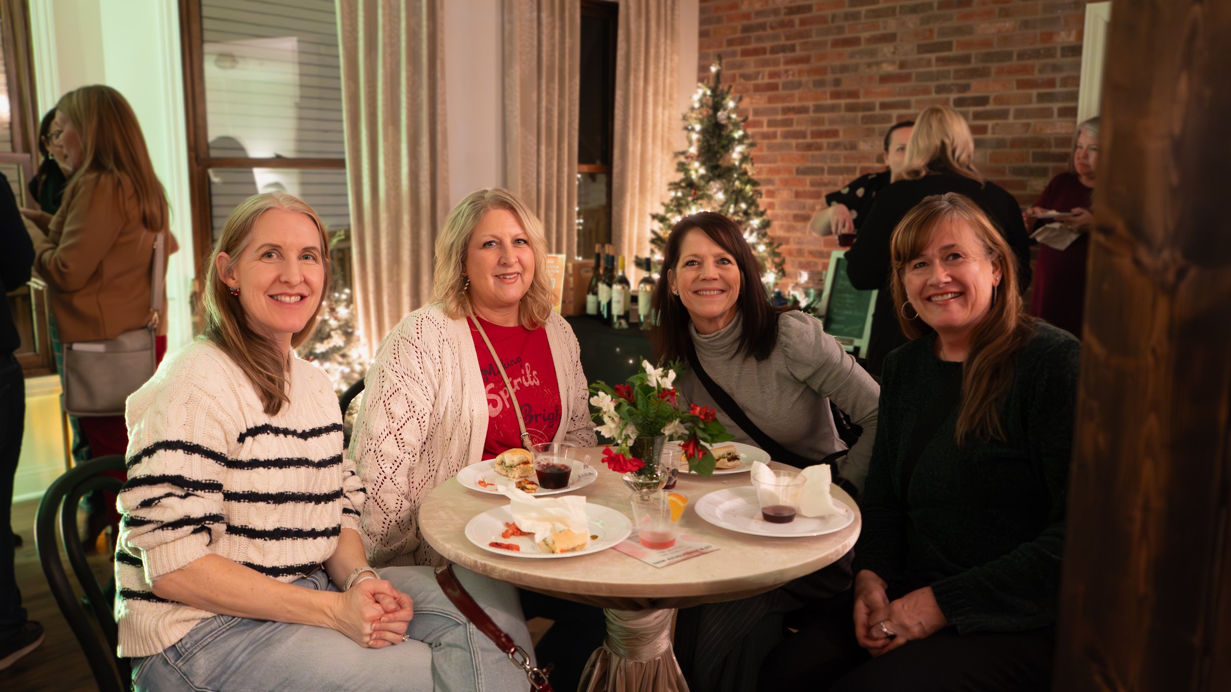Four women sitting at a table decorated with a small Christmas tree and poinsettia in a festive setting with other people in the background.