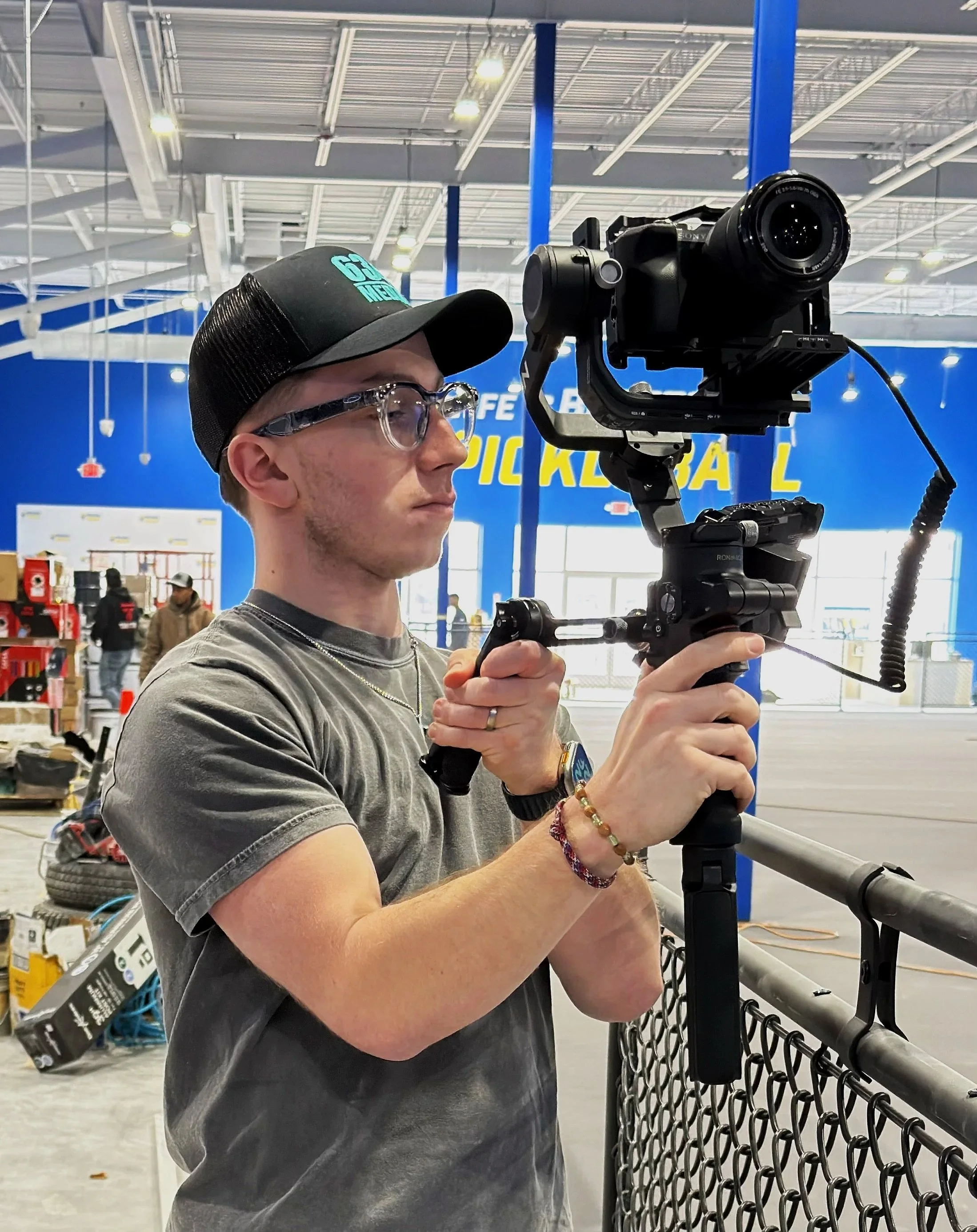 A young man with glasses, a hat, and jewelry is operating a camera mounted on a stabilizer rig at a retail store with a large blue and yellow sign in the background.