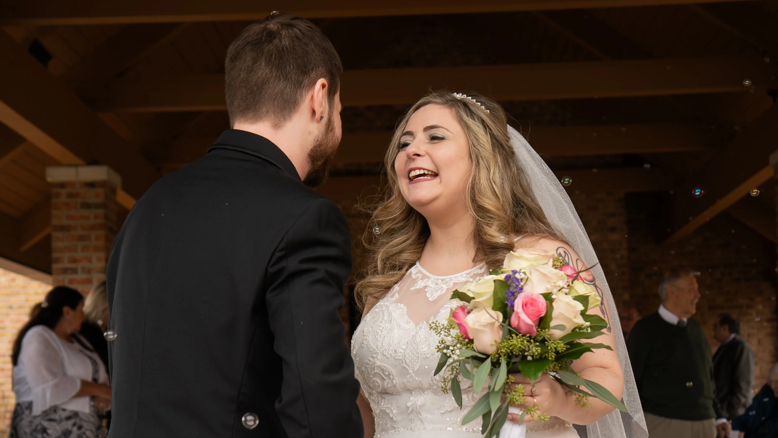 A bride and groom on their wedding day, smiling at each other indoors with brick walls and wooden beams.