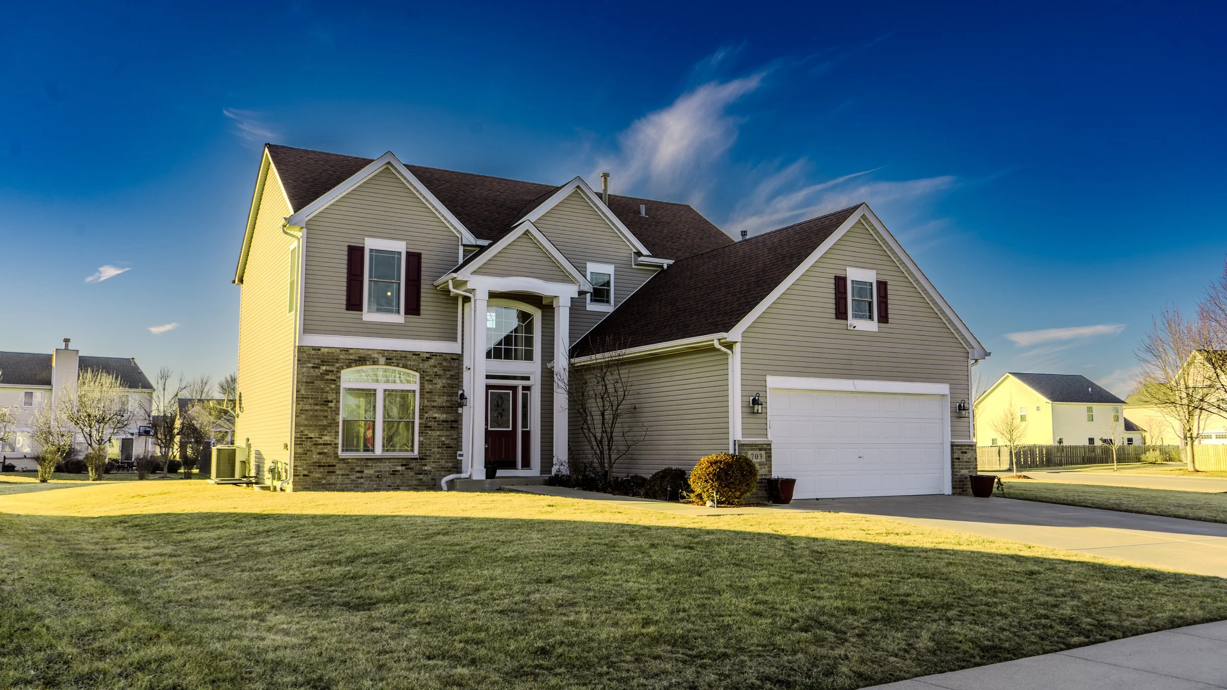 Front view of a two-story suburban house with a garage, gray siding, stone accents, a small front porch, and a well-maintained lawn under a partly cloudy blue sky.