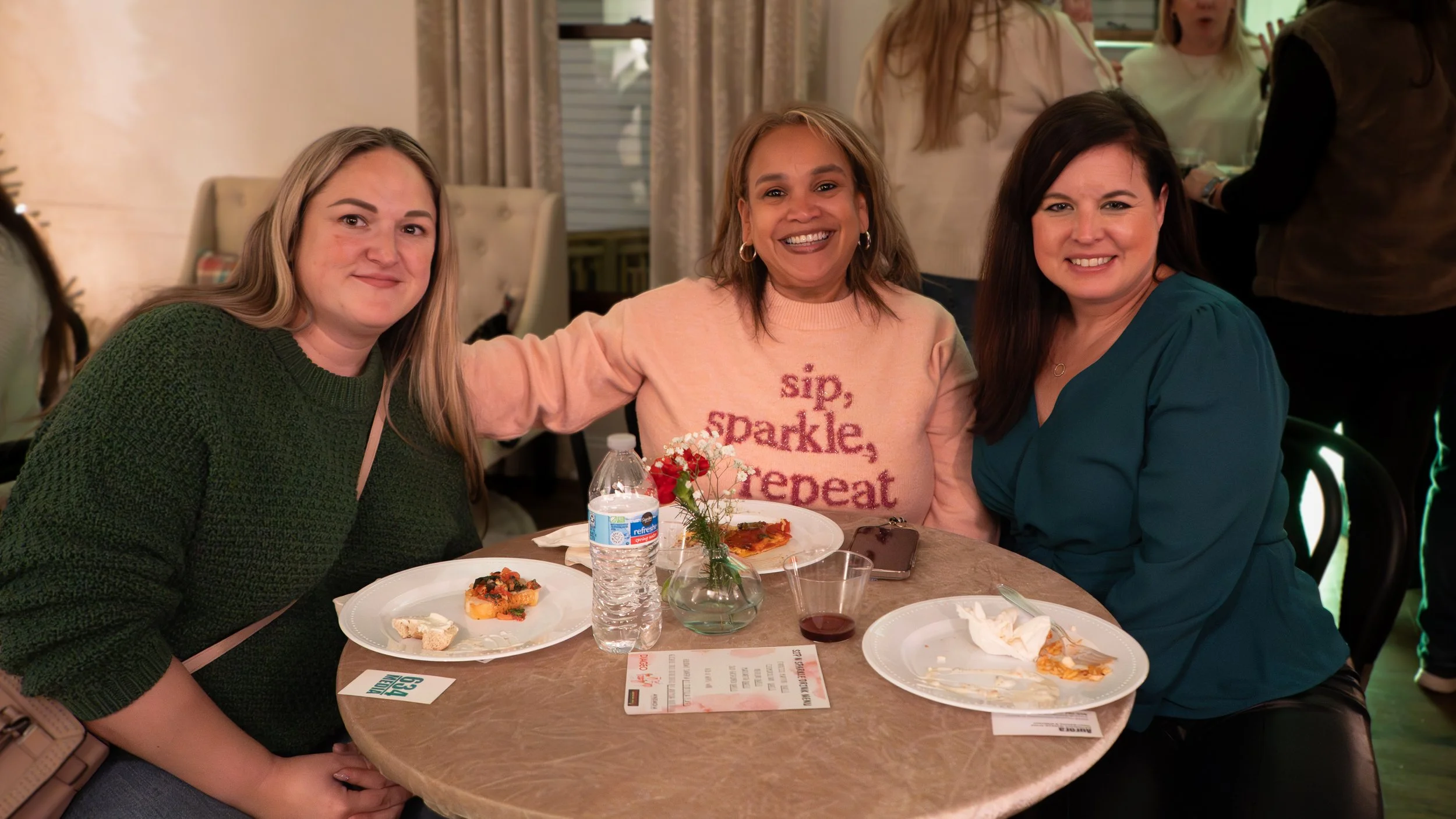 Three women sitting at a dining table smiling for the camera with two plates of food, a water bottle, a glass of wine, and a small flower arrangement in the center.