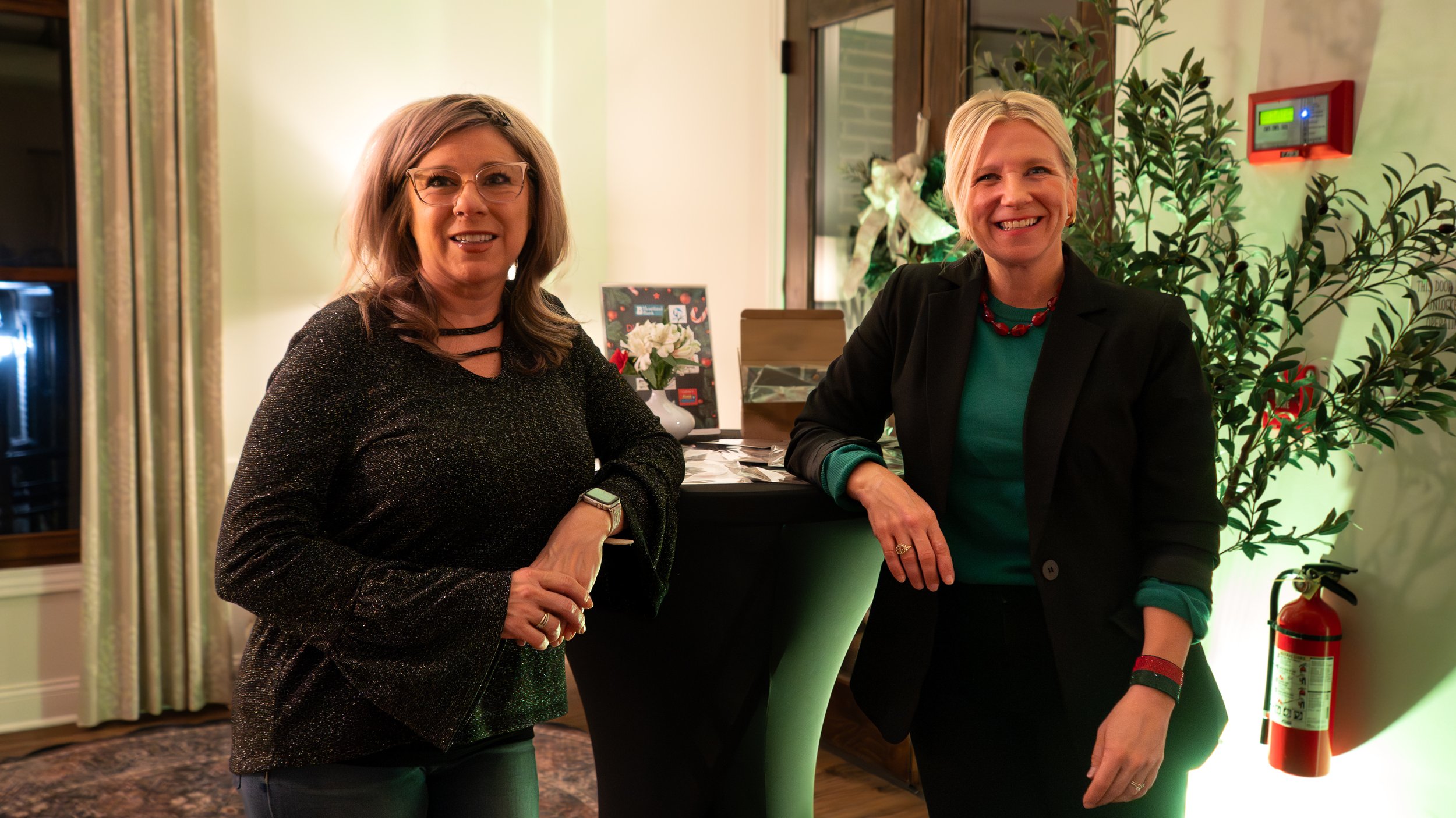 Two women standing and smiling indoors next to a counter decorated with flowers; one has glasses and brown hair, the other has short blonde hair; background includes a window and a fire extinguisher.