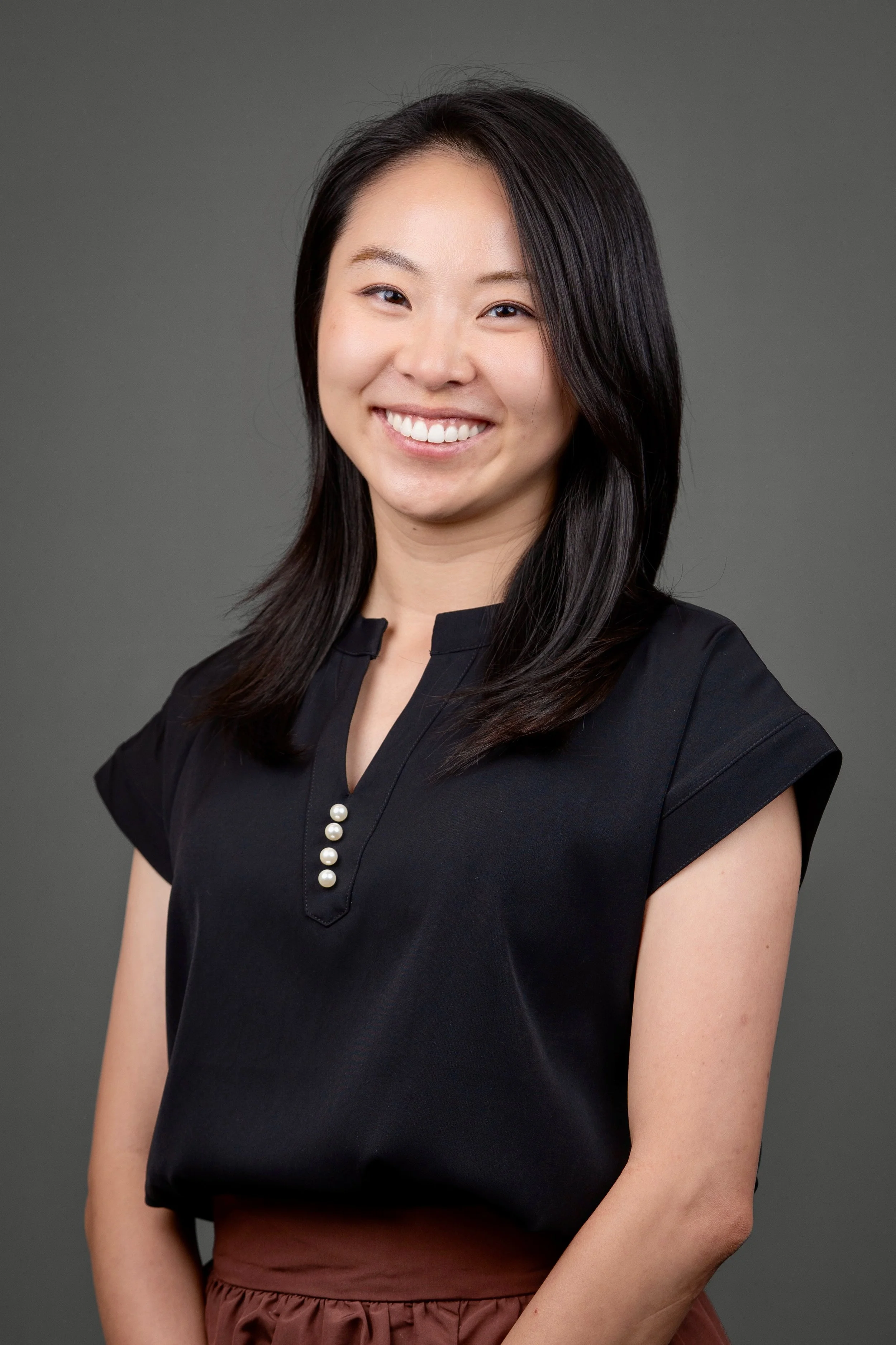 A young woman with straight black hair, smiling, wearing a black top with pearl buttons and brown skirt, against a gray background.
