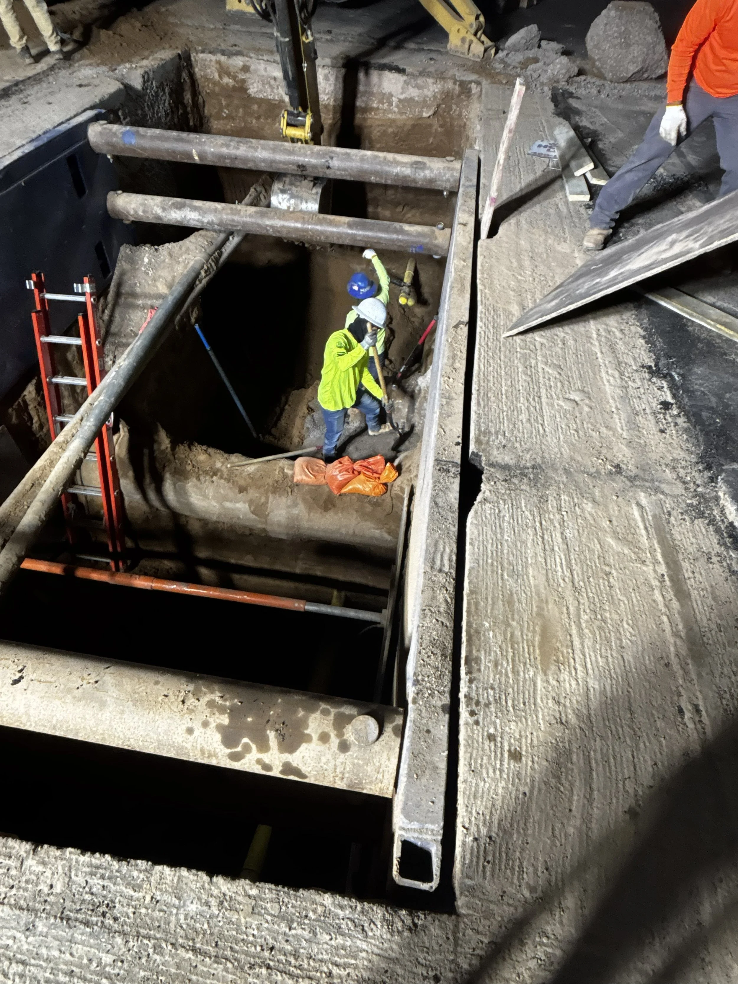 Construction workers in safety gear working inside a large rectangular hole in a construction site, surrounded by scaffolding and equipment.