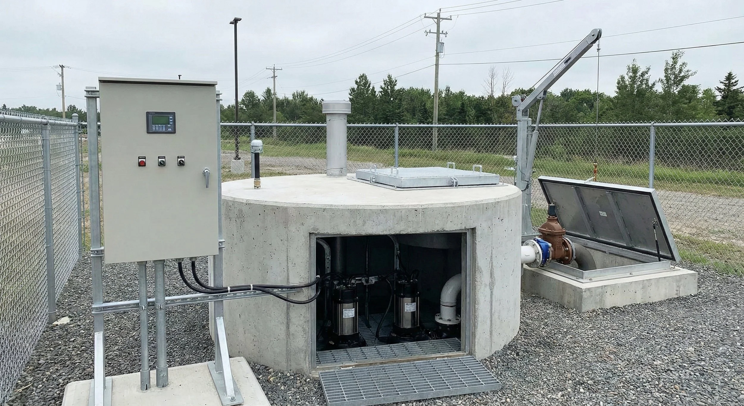 An underground water pump station with control panel, pumps, pipes, and a retractable cover, surrounded by a wire fence and utility poles in the background.